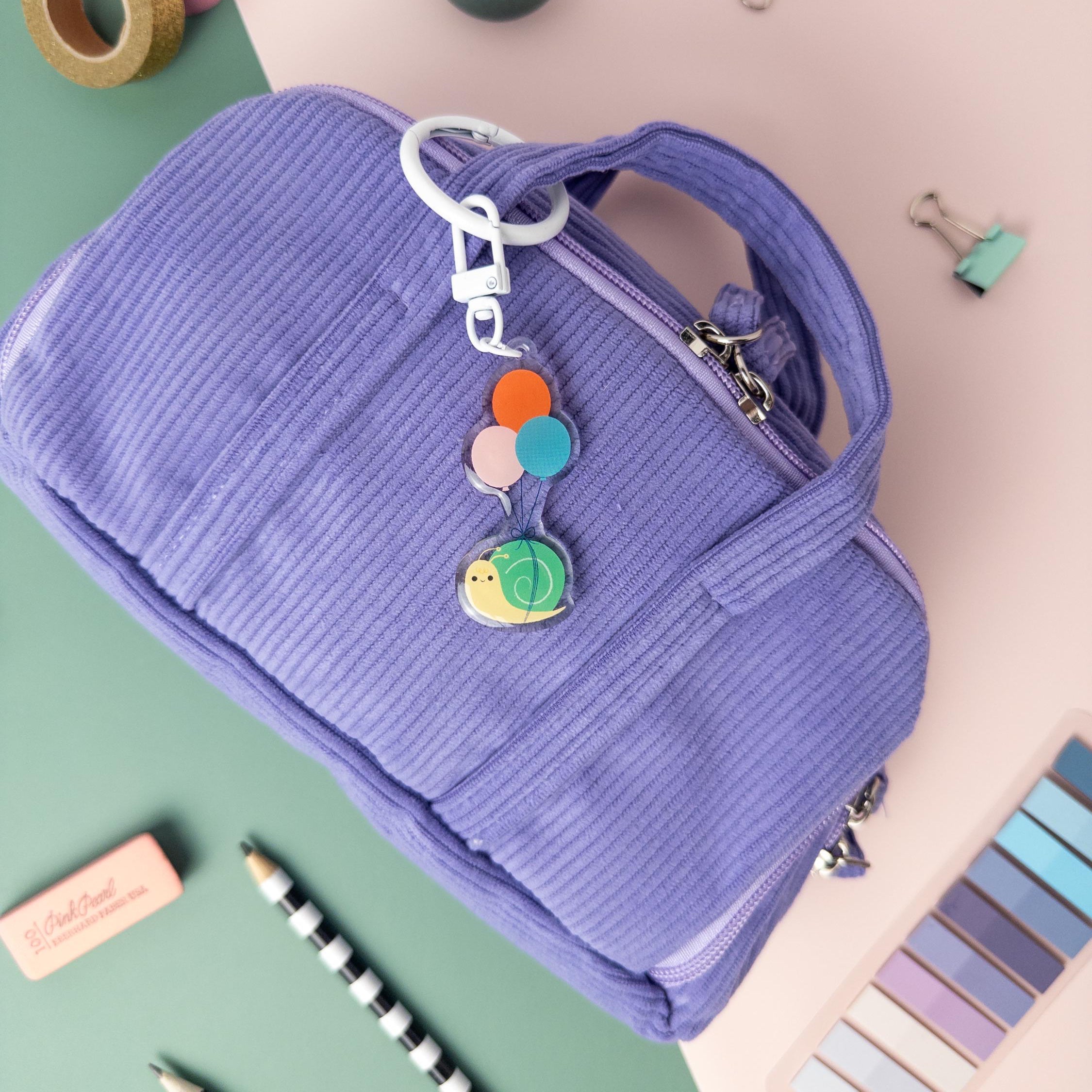 Colorful acrylic keychain featuring a snail being carried by balloons. The keychain is clipped to a small textured purple bag photographed on a pink and teal background.