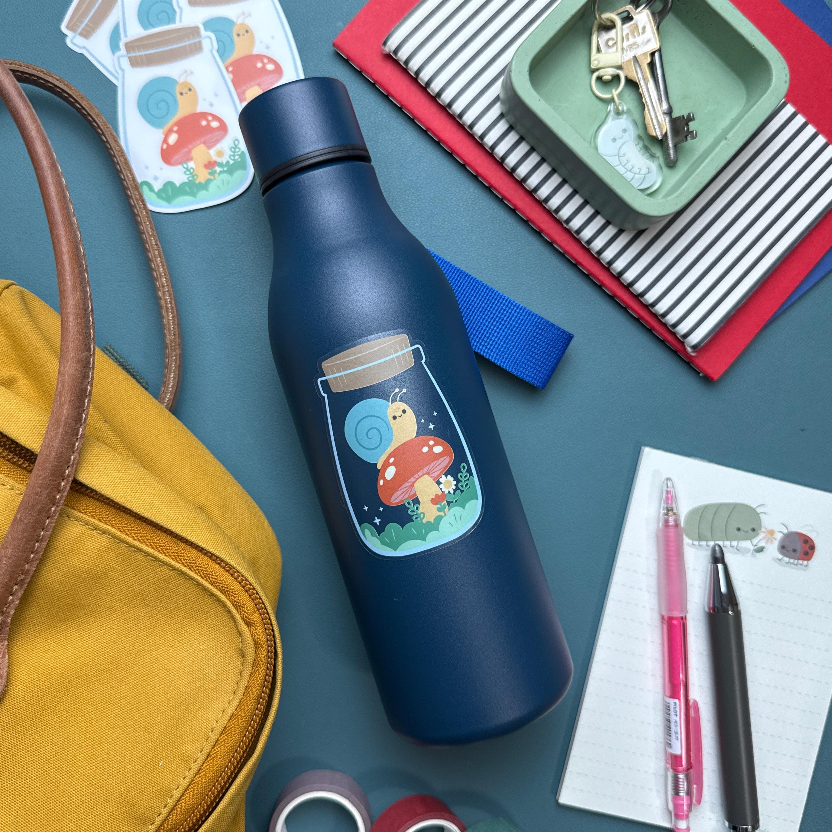 Illustrated sticker of a terrarium jar with snail sitting atop a mushroom. It is pictured attached to a dark blue water bottle and is surrounded by a stack of similar stickers, a bag, and other art supplies.