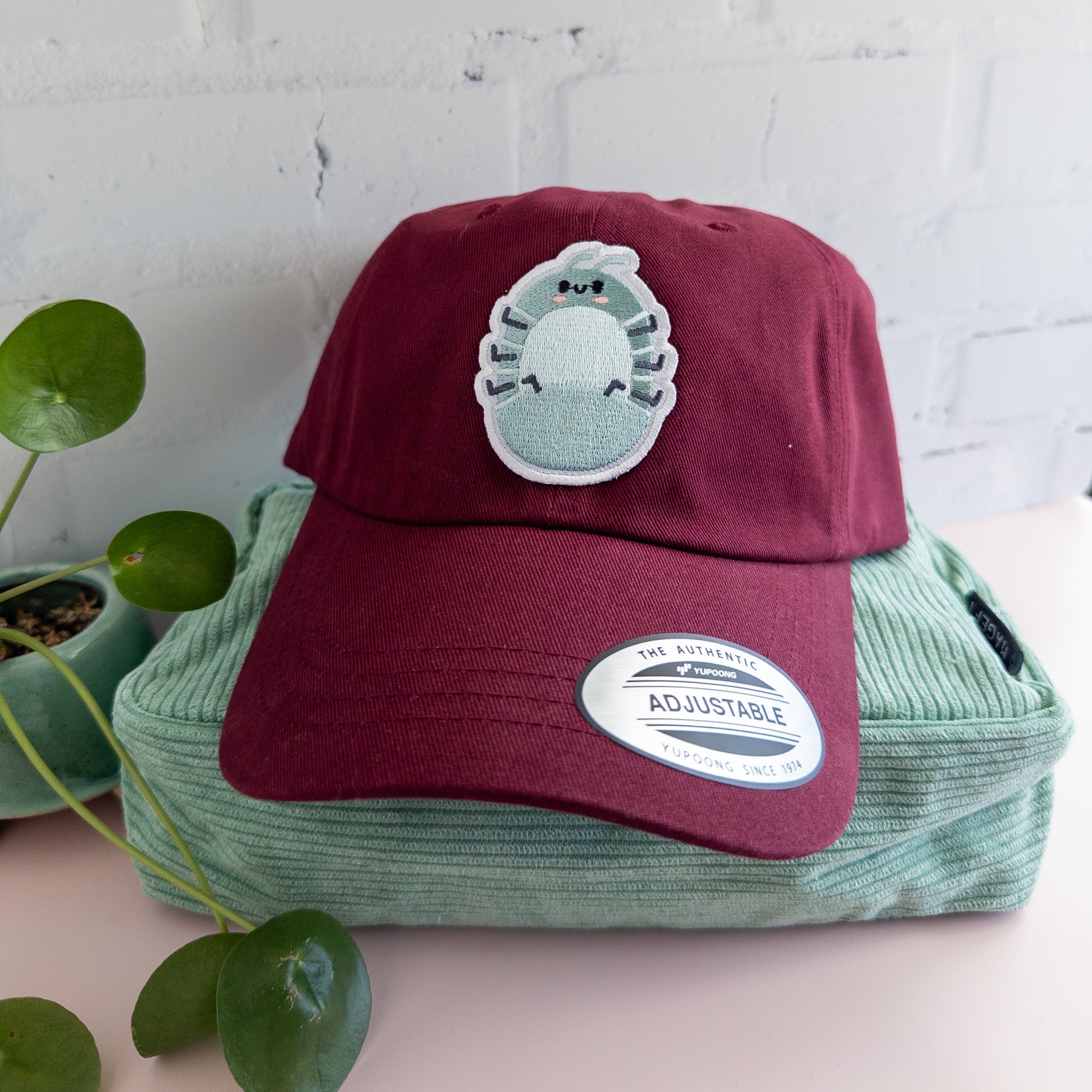 Maroon hat with teal roly poly patch on front sitting atop a mint green corduroy bag with plant sitting beside it.