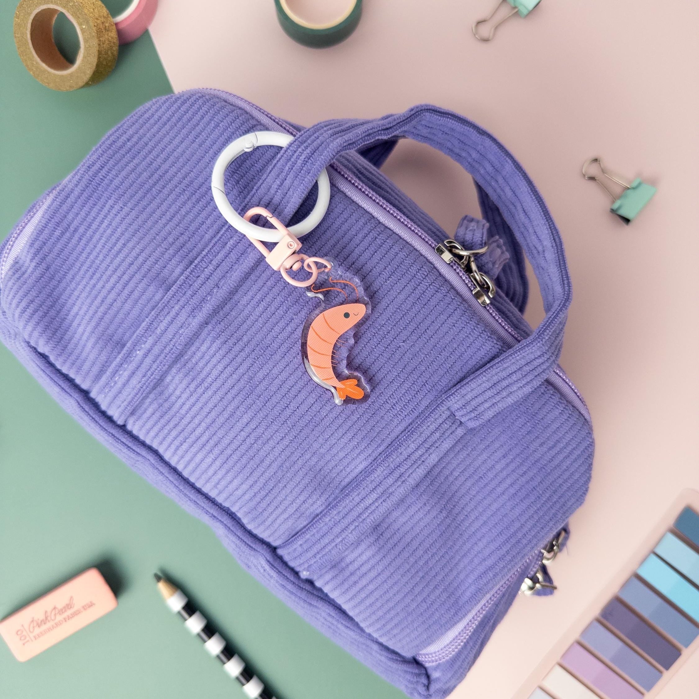 Pink shrimp acrylic charm keychain attached to a purple textured bag with a variety of office supplies surrounding it.