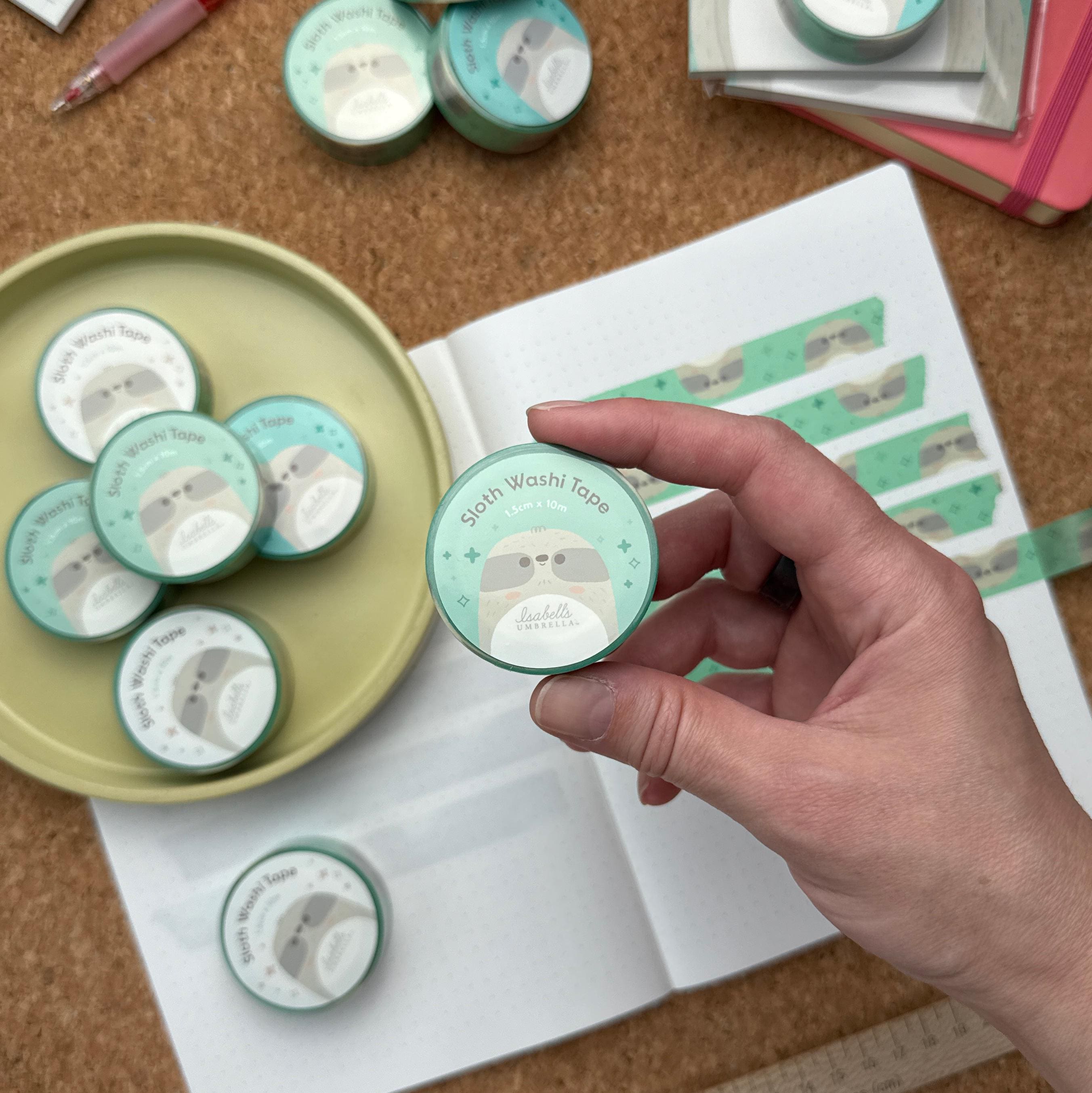 Hand holding a roll of sloth themed washi tape. It is pictured on a cork textured background with rolls of washi tape and an open notebook with strips of sloth washi tape taped across the page.