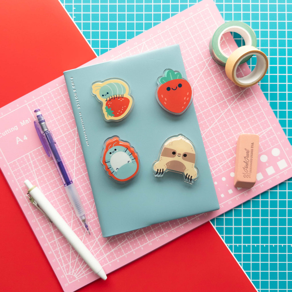 Four cute acrylic journal clips featuring a roly poly, roly poly on a strawberry, sloth, and strawberry on a notebook with stationery supplies.