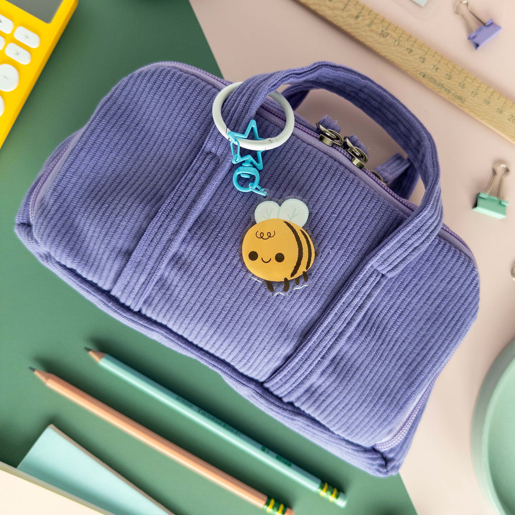 Purple corduroy pencil case with a bee-themed keychain on a school supplies background