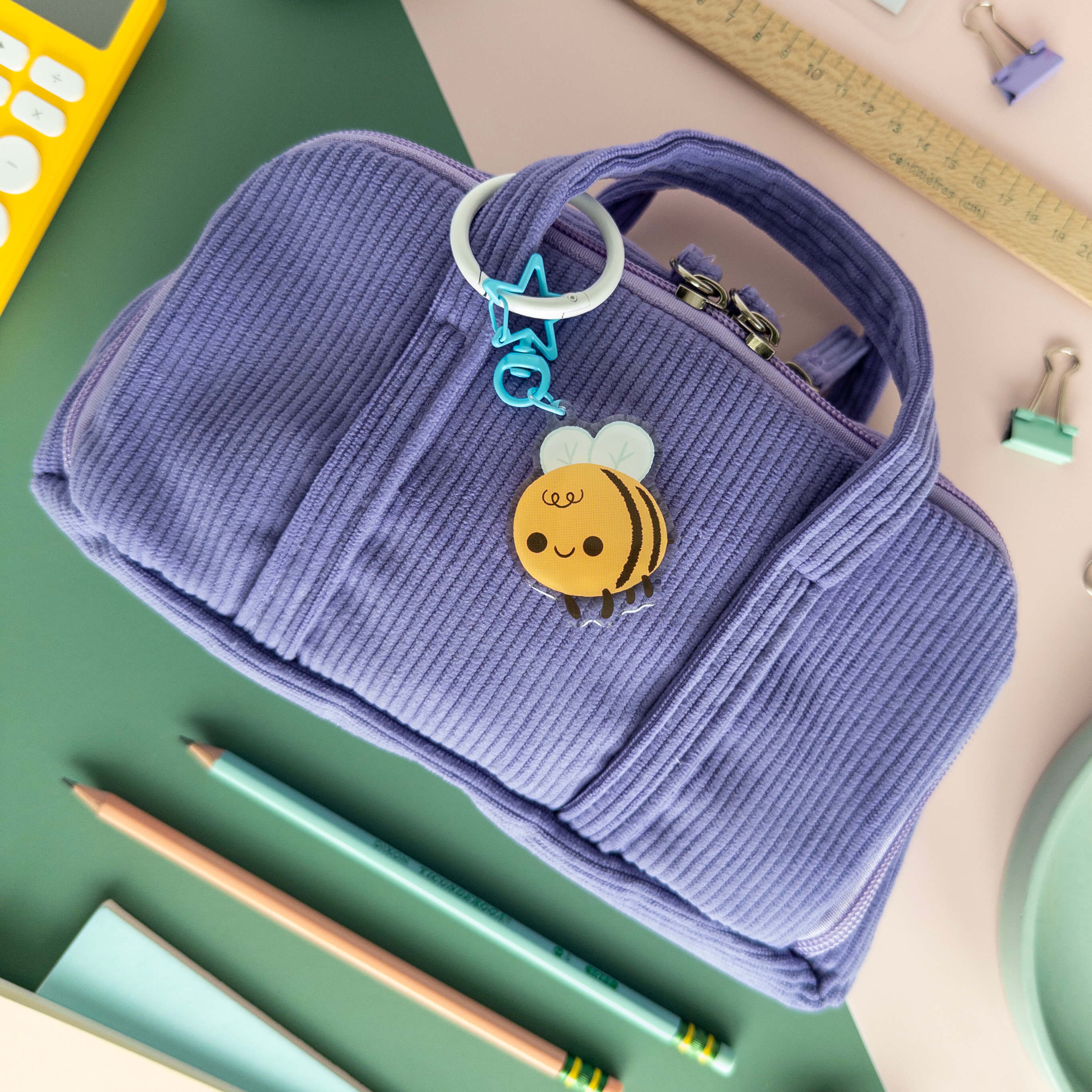 Purple corduroy pencil case with a bee-themed keychain on a school supplies background