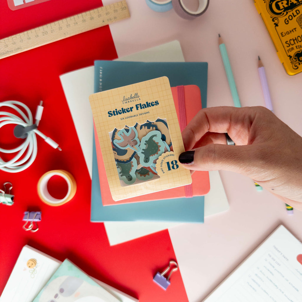 A hand holds a pack of cute dinosaur sticker flakes over colorful notebooks and stationery.