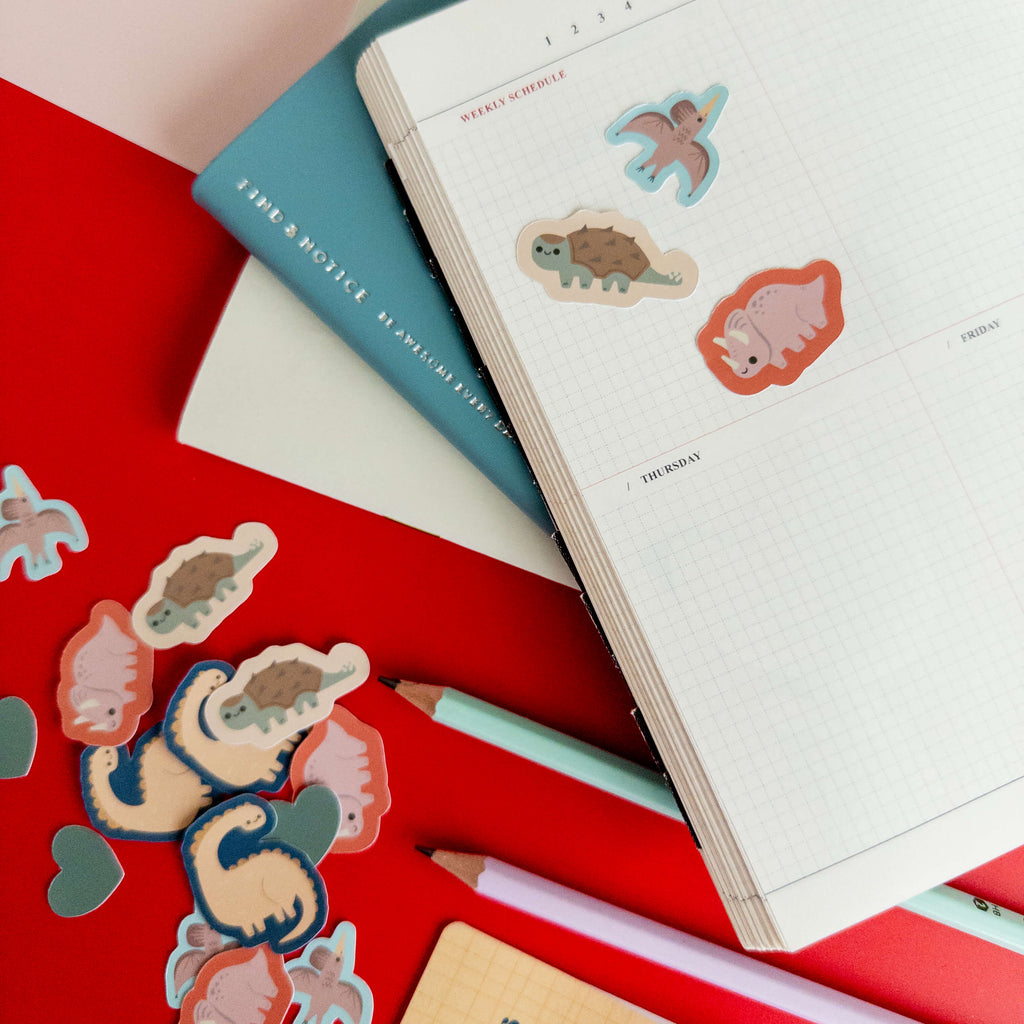 Cute dinosaur stickers decorating an open grid notebook surrounded by colorful stationery.