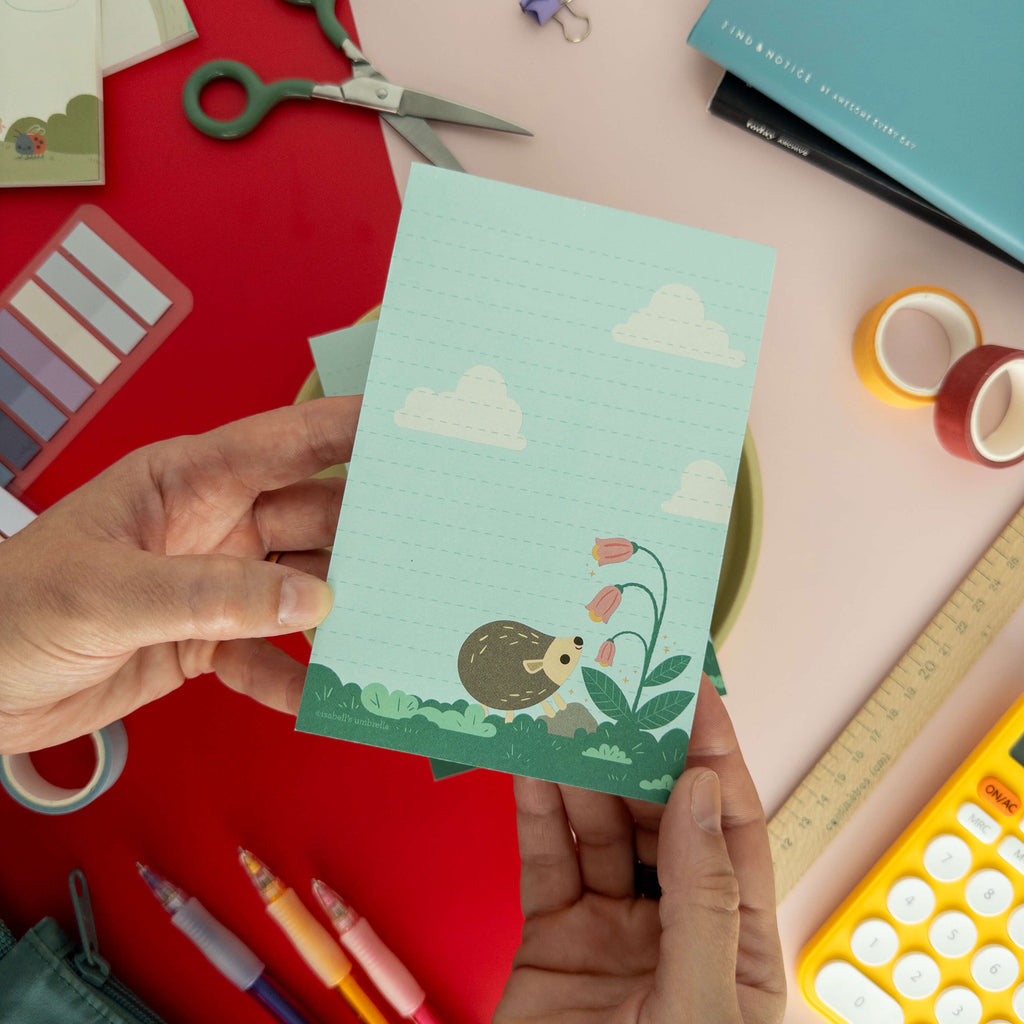 A hand holds a cute hedgehog illustrated notepad surrounded by colorful stationery supplies.