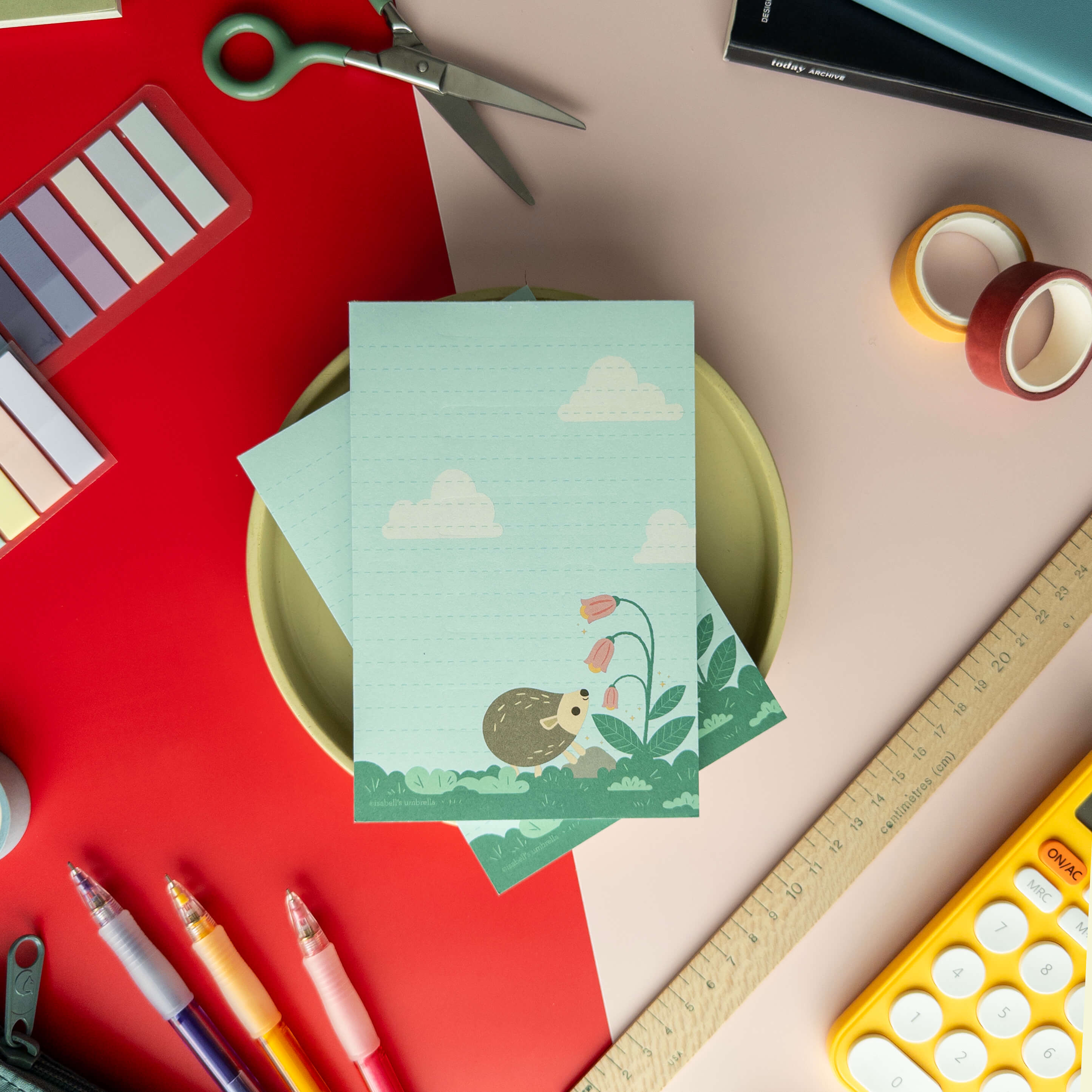 Cute hedgehog notepad displayed on a colorful desk with pens, scissors, tape, and stationery supplies.