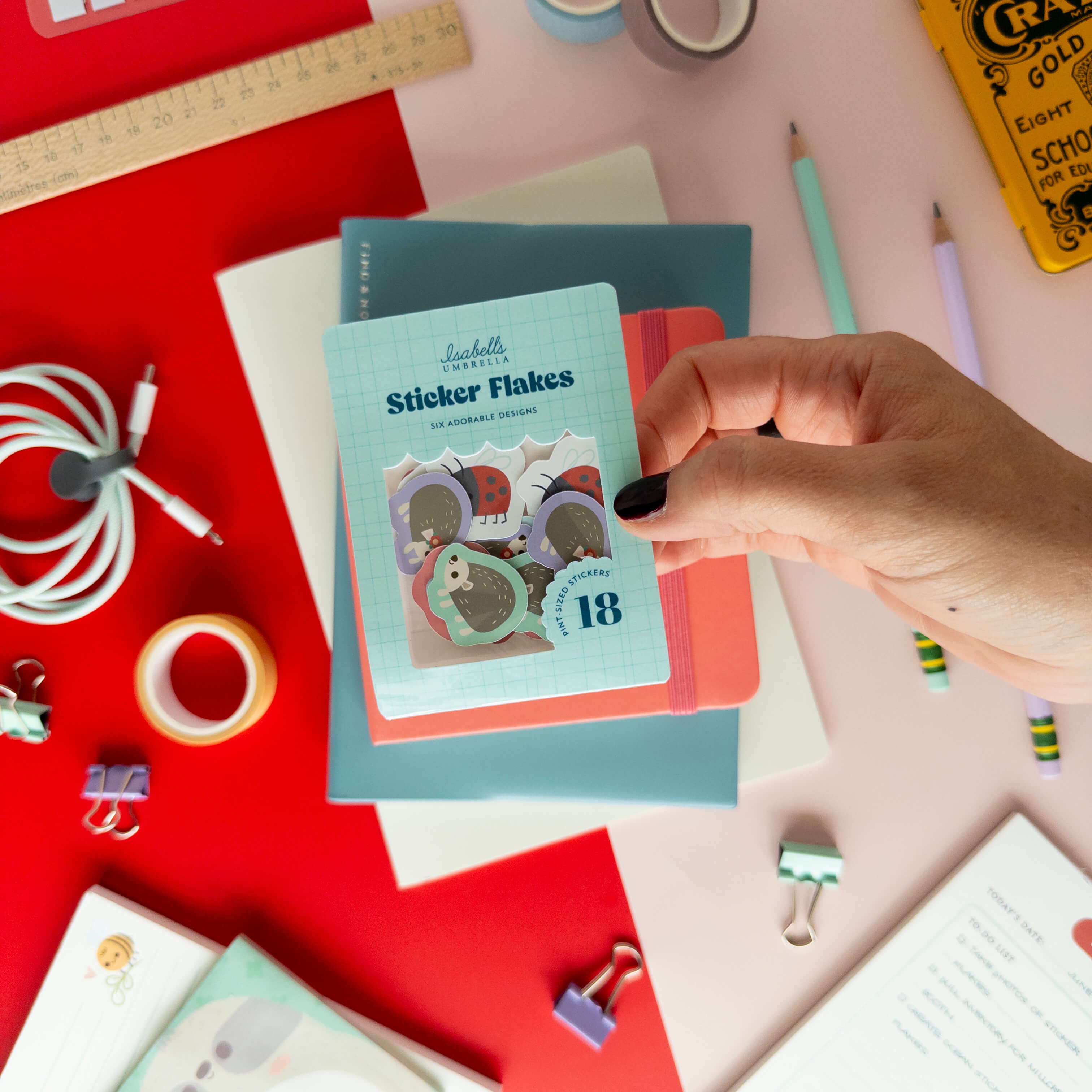 A hand holds a pack of cute hedgehog sticker flakes over colorful notebooks and stationery.