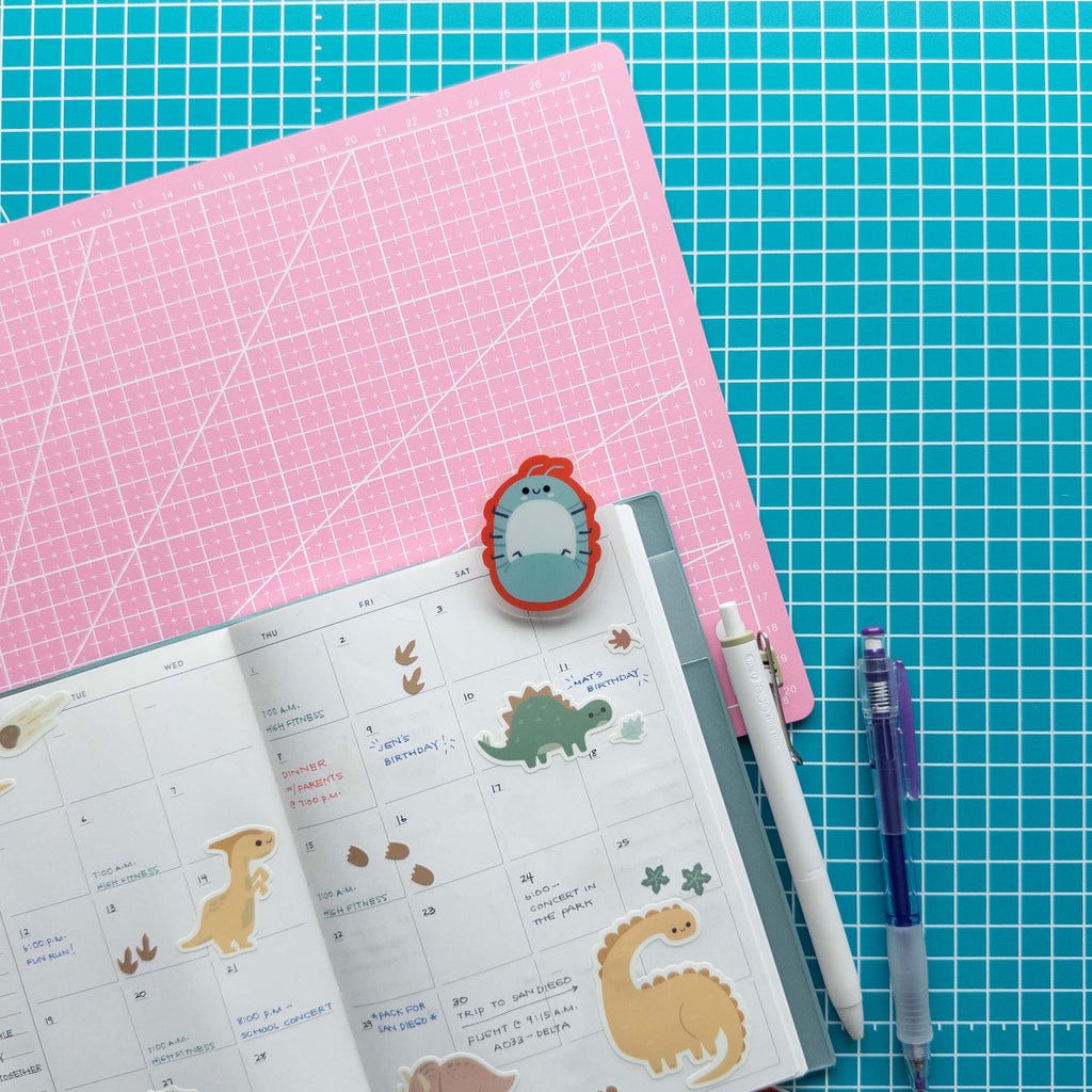 Cute roly poly acrylic journal clip attached to an open planner surrounded by colorful stationery supplies.