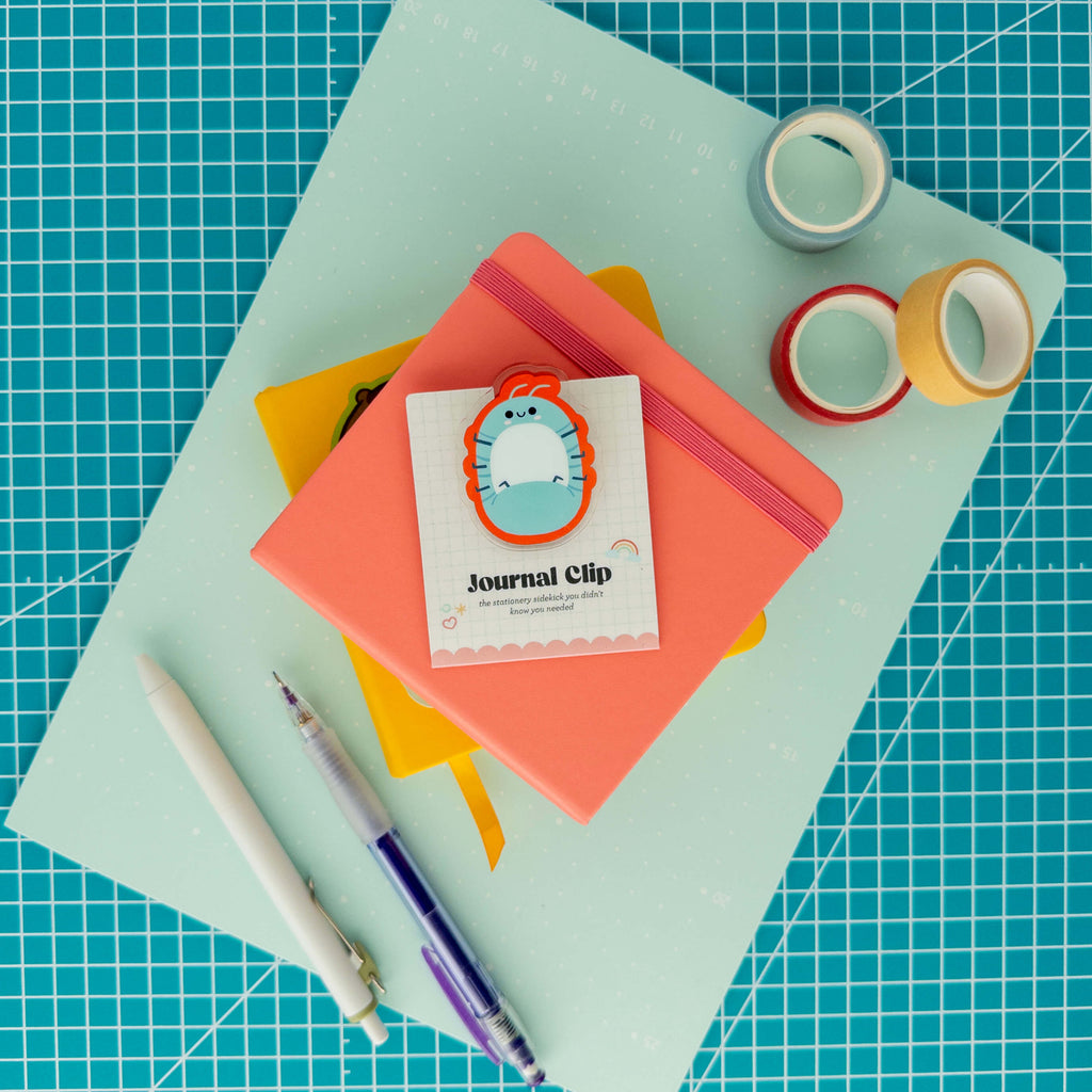 Cute roly poly acrylic journal clip displayed on its backing card with colorful paper, washi tape, and pens.