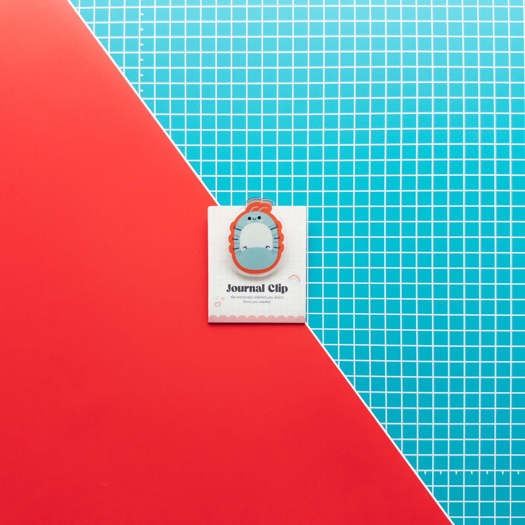 Cute roly poly acrylic journal clip displayed on its branded backing card against a colorful red and grid background.