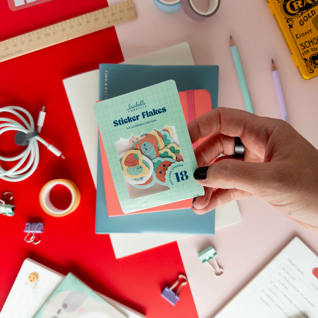 A hand holds a pack of cute roly poly sticker flakes over colorful notebooks and stationery.