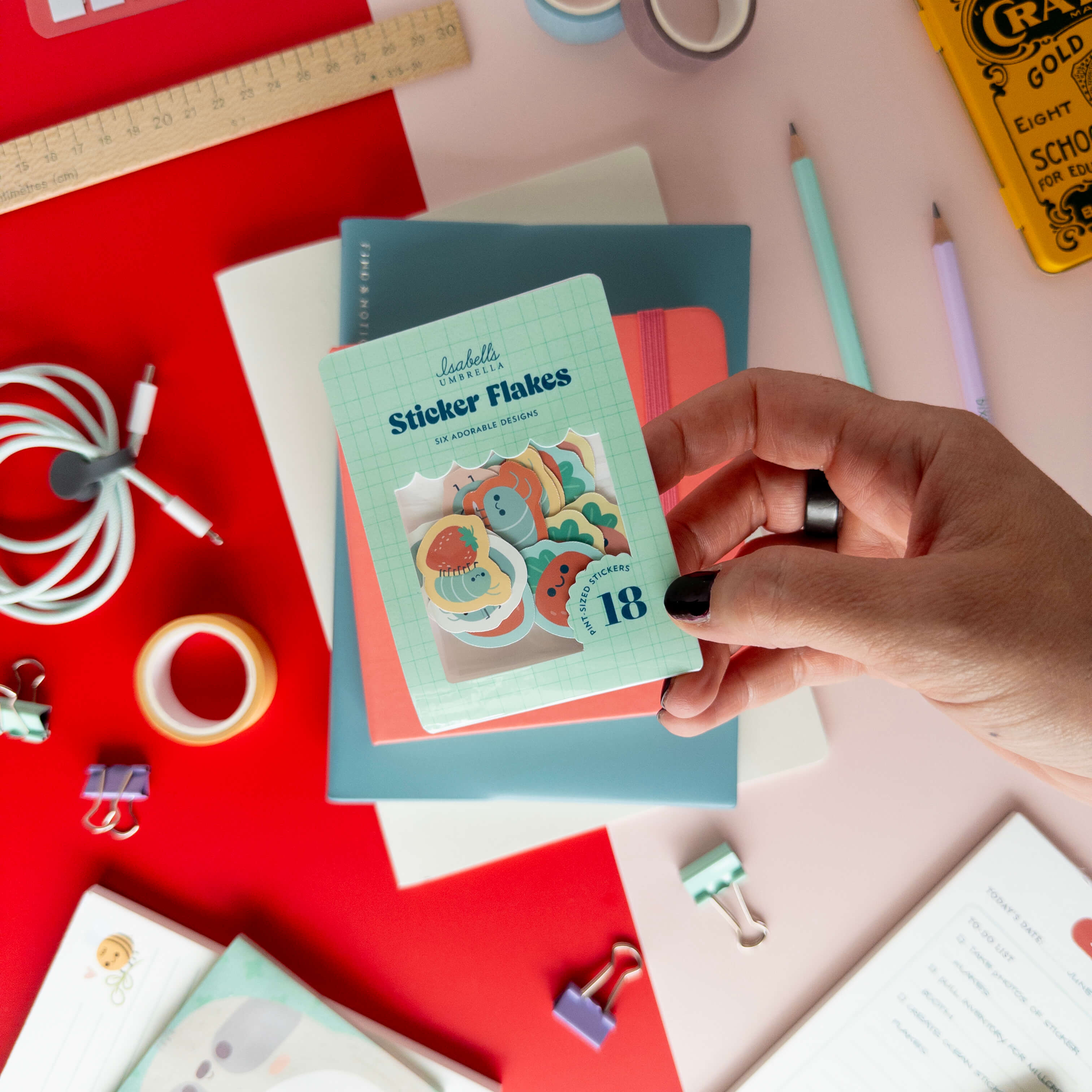 A hand holds a pack of cute roly poly sticker flakes over colorful notebooks and stationery.