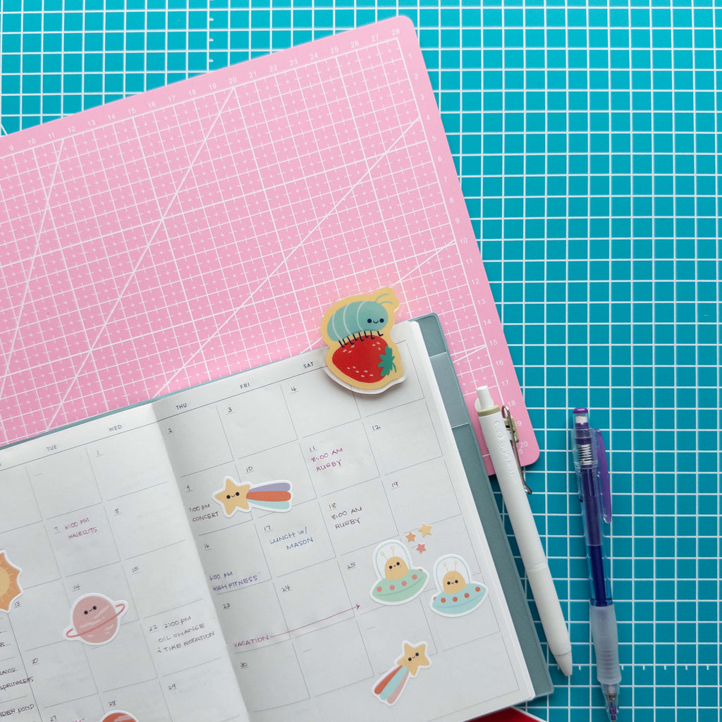 Cute roly poly on a strawberry acrylic journal clip attached to an open planner with pens and stationery.