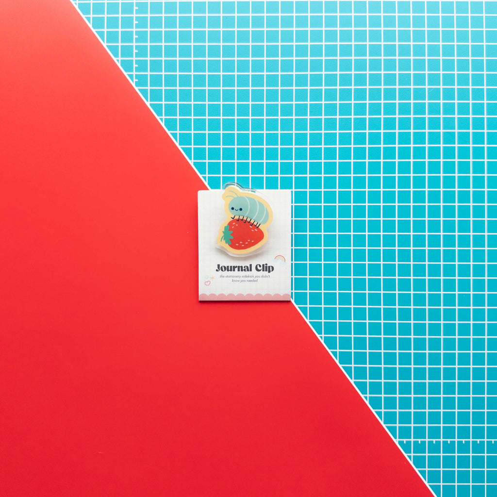 Cute roly poly on a strawberry acrylic journal clip displayed on its branded backing card against a colorful red and grid background.