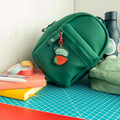 Cute roly poly on a strawberry acrylic keychain hanging from a green backpack with stationery supplies nearby.