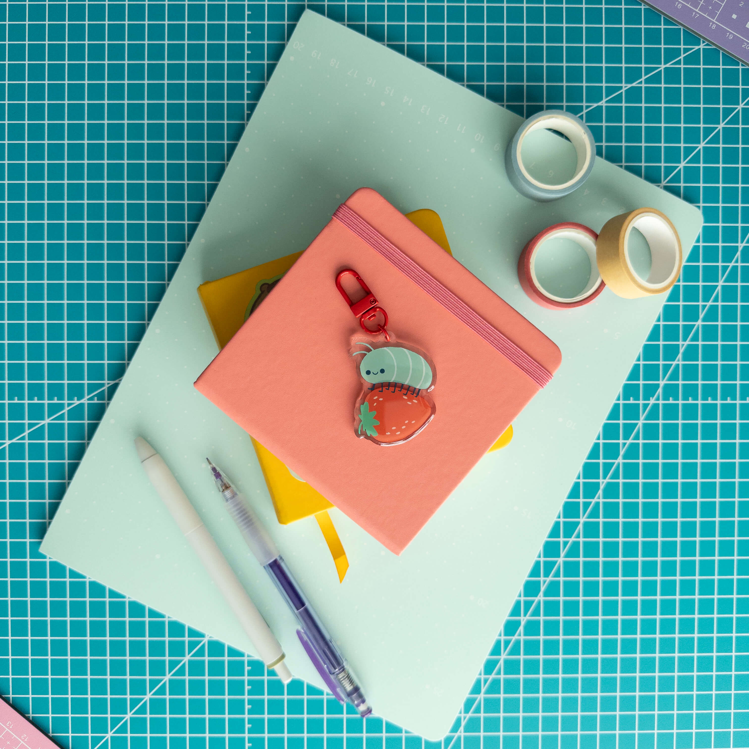 Cute roly poly on a strawberry acrylic keychain displayed with colorful notebooks, paper, and stationery.