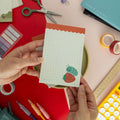 A hand holds a cute roly poly on a strawberry illustrated notepad surrounded by colorful stationery.