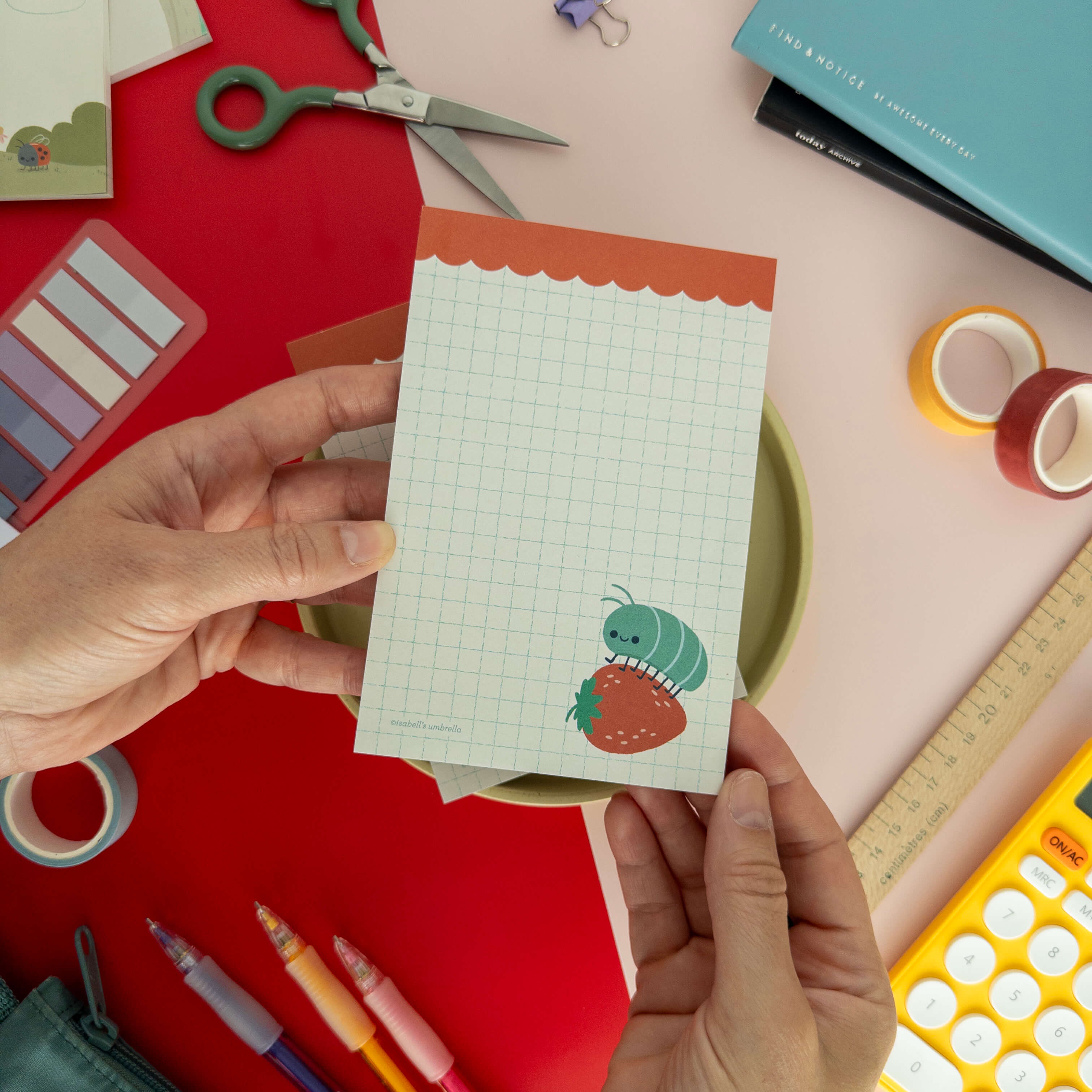 A hand holds a cute roly poly on a strawberry illustrated notepad surrounded by colorful stationery.