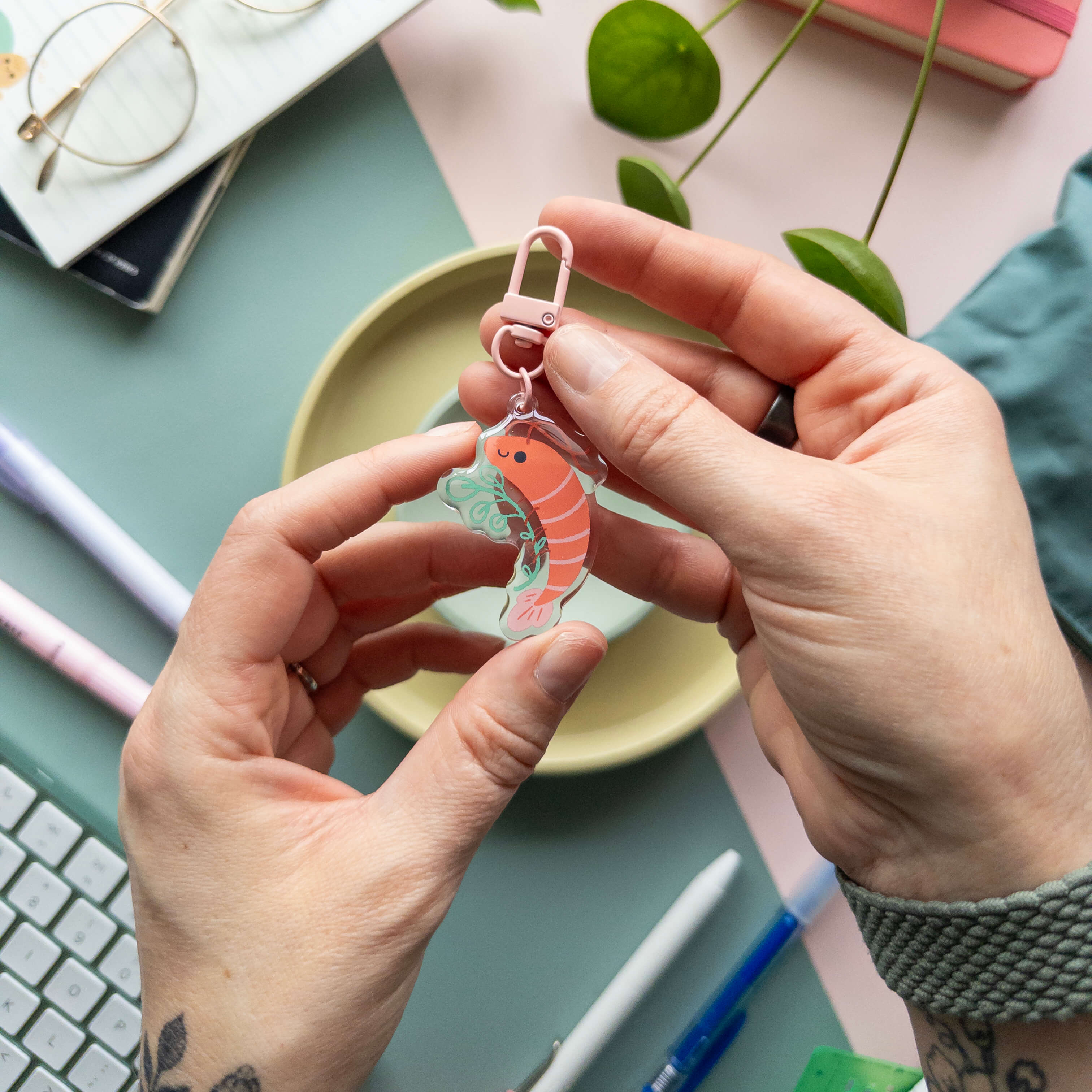Person holding a keychain with a shrimp design over a desk with stationery items.