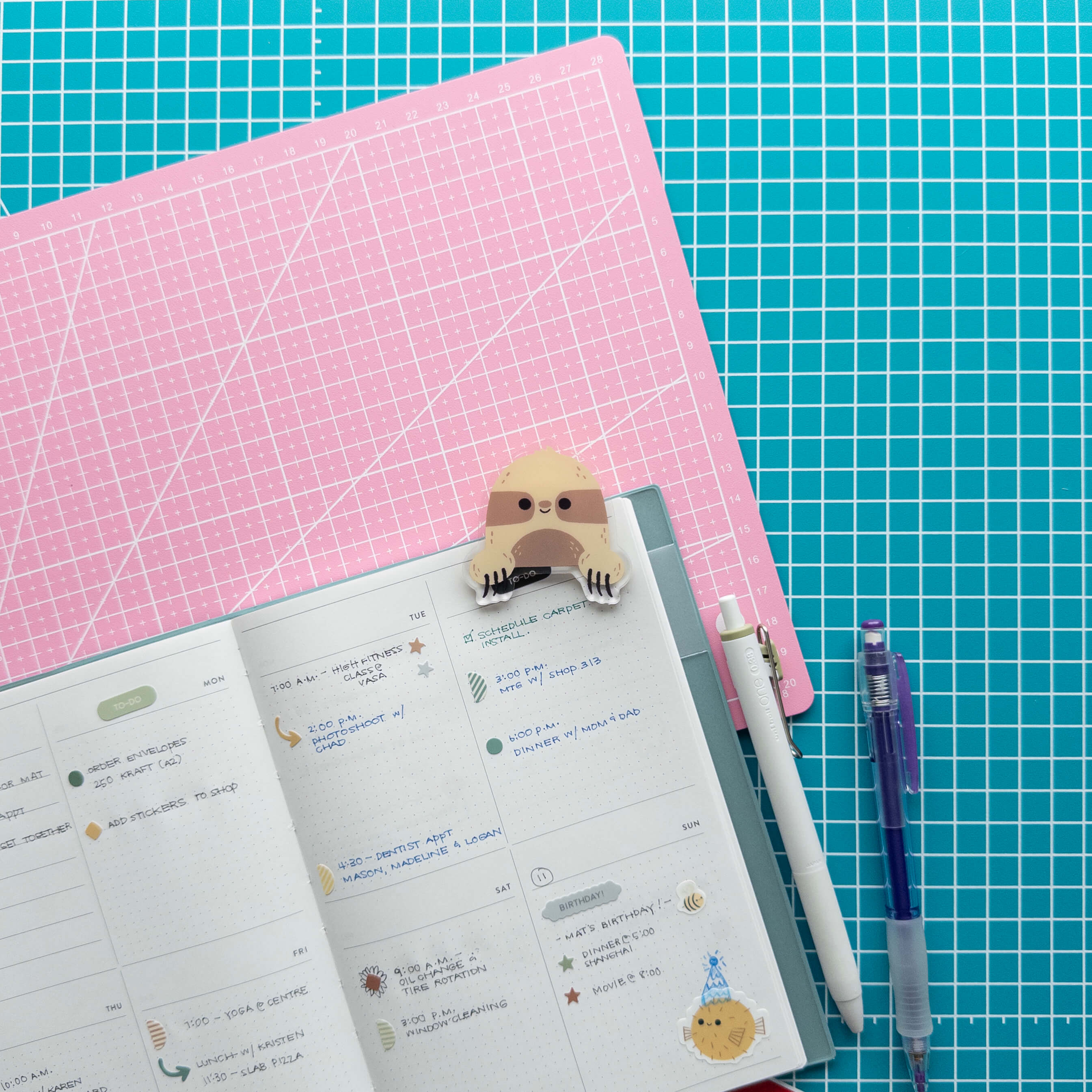 Cute sloth acrylic journal clip attached to an open planner with pens and stationery supplies.