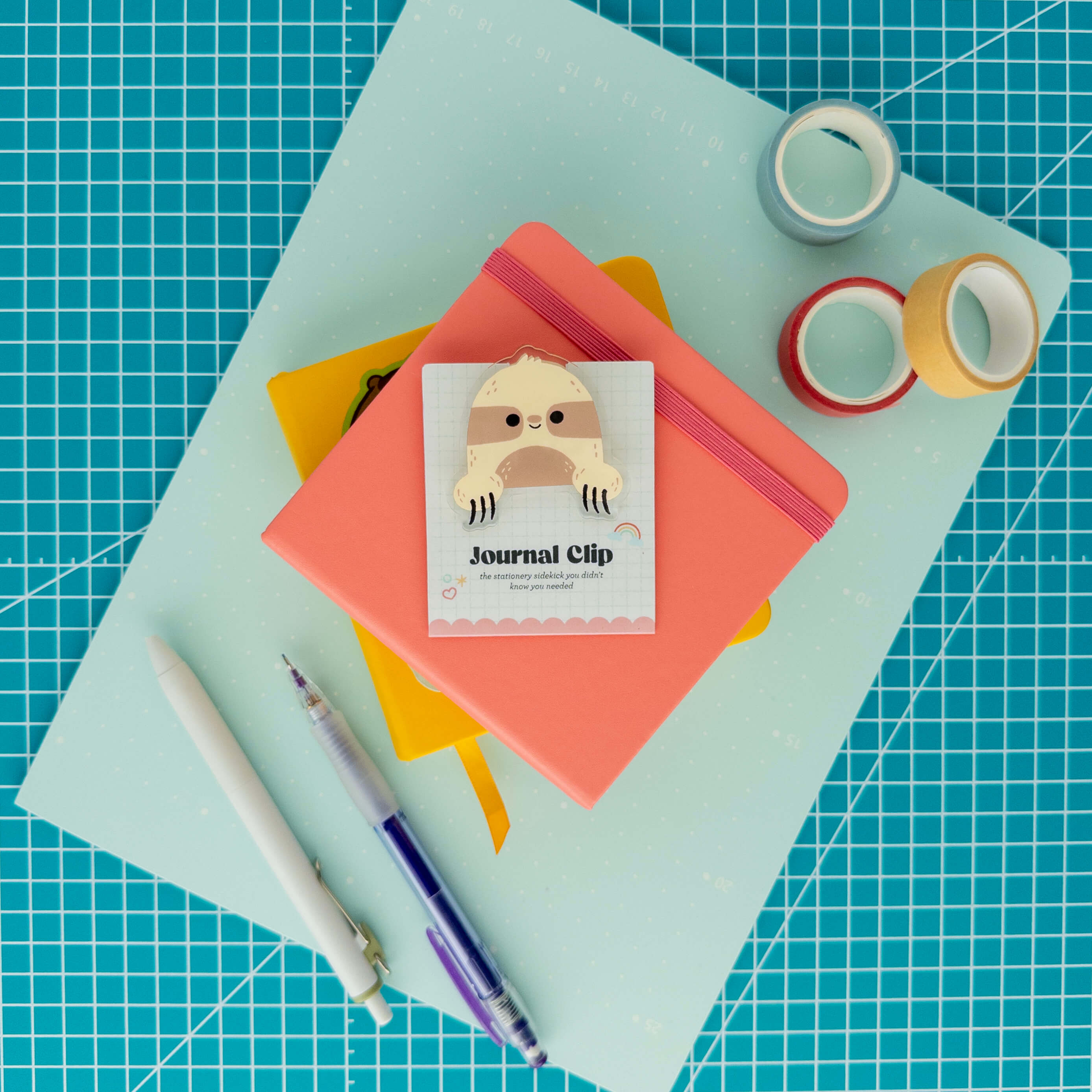 Cute sloth acrylic journal clip displayed on its backing card with colorful paper, washi tape, and pens.