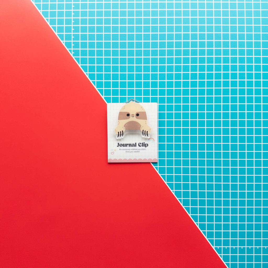 Cute sloth acrylic journal clip displayed on its branded backing card against a colorful red and grid background.