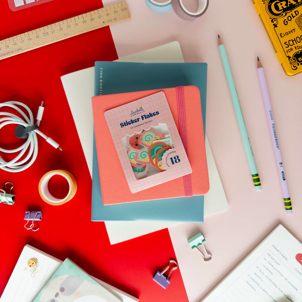 A hand holds a pack of cute snail sticker flakes over colorful notebooks and stationery.