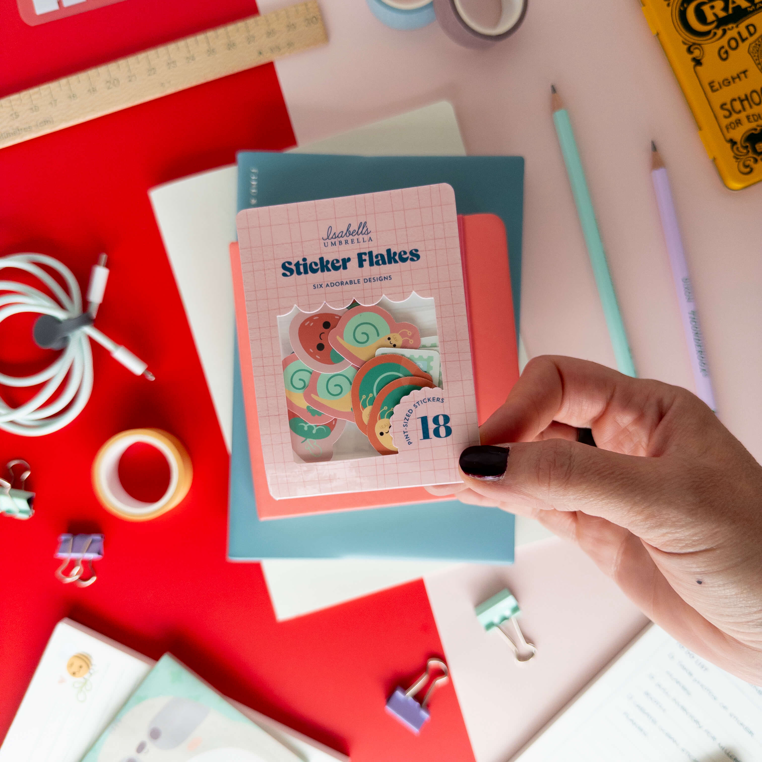 A hand holds a pack of cute snail sticker flakes over colorful notebooks and stationery.