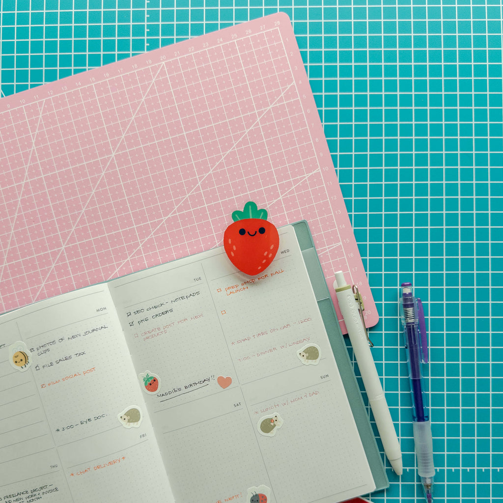 Cute strawberry acrylic journal clip attached to an open planner with pens and stationery supplies.