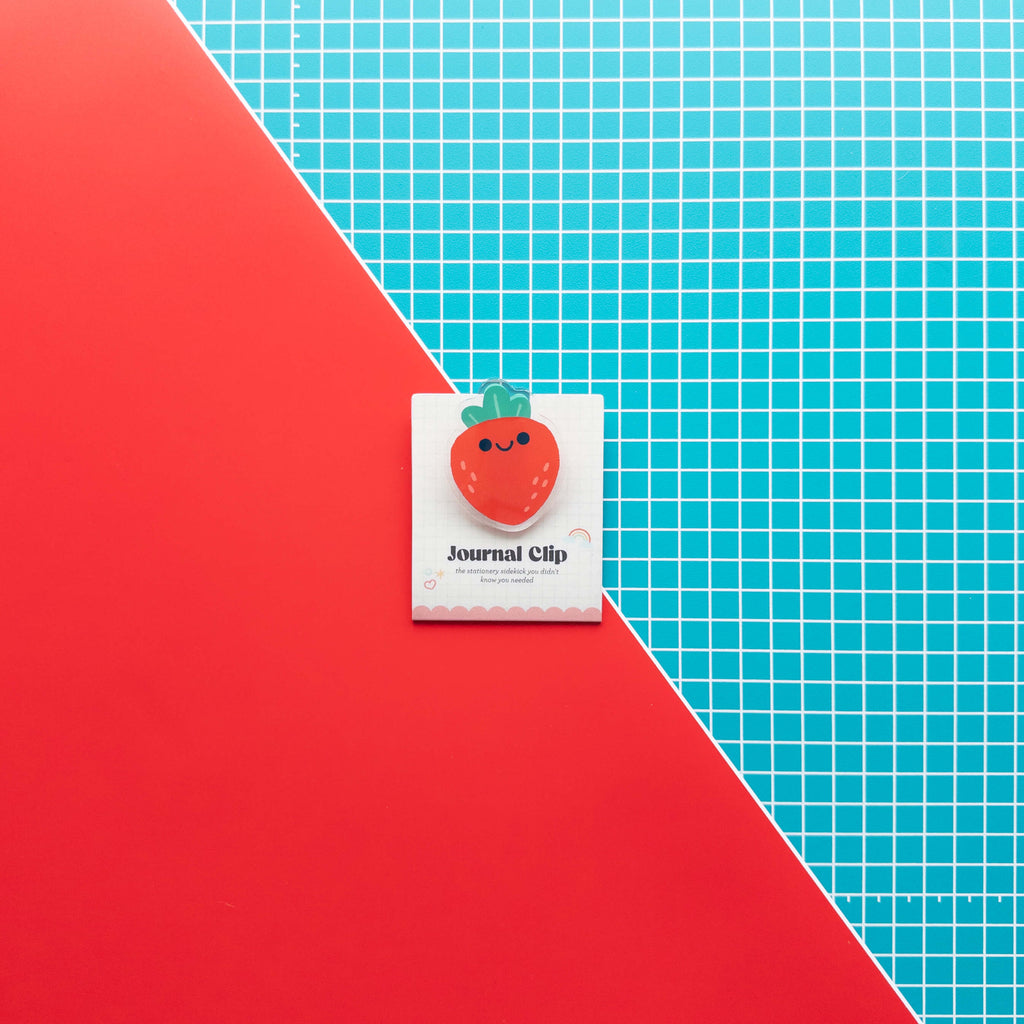 Cute strawberry acrylic journal clip displayed on its branded backing card against a colorful red and grid background.