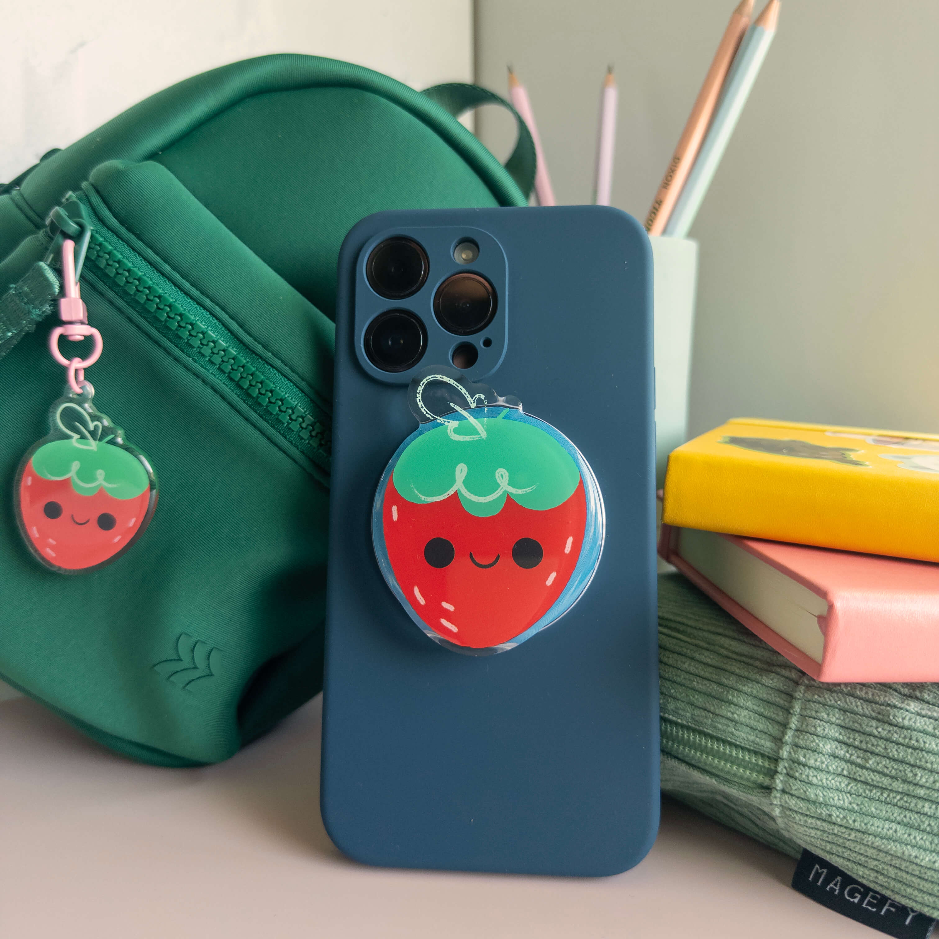Blue phone case with a strawberry magnetic phone grip on a desk with a green bag with a strawberry keychain and books.