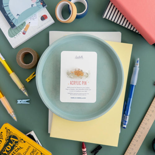 Adorable acrylic pin featuring a chubby ankylosaurus made from durable acrylic and finished with a smooth epoxy finish. It is set on a blue plate that is surrounded by a variety of art supplies. 