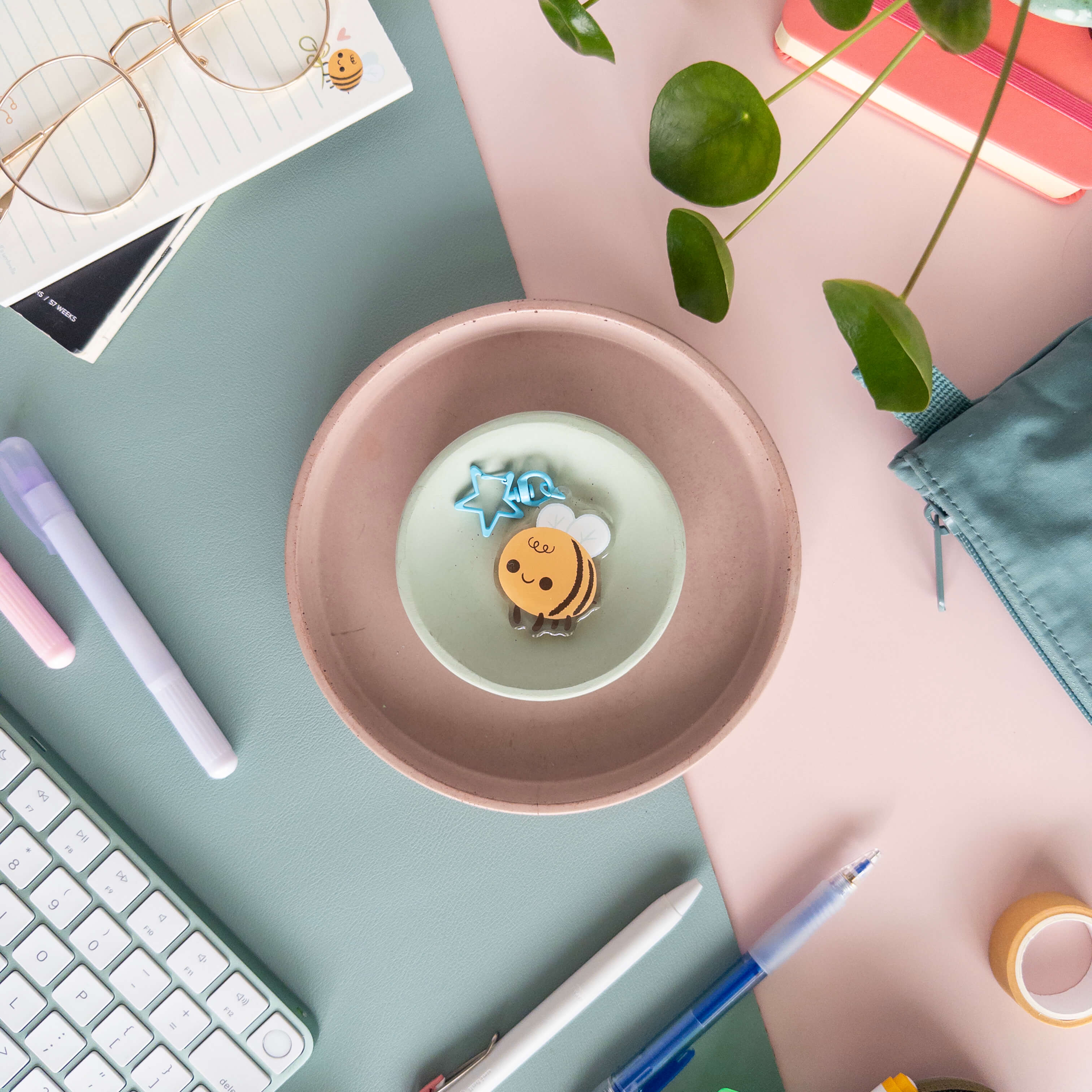 Desk setup with a pink plate containing a small bee charm keychain pictured on a pink and teal background surrounded by a variety of stationery supplies.