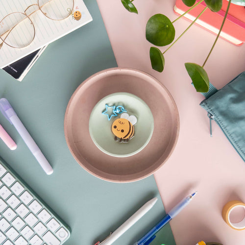 Desk setup with a pink plate containing a small bee charm keychain pictured on a pink and teal background surrounded by a variety of stationery supplies.