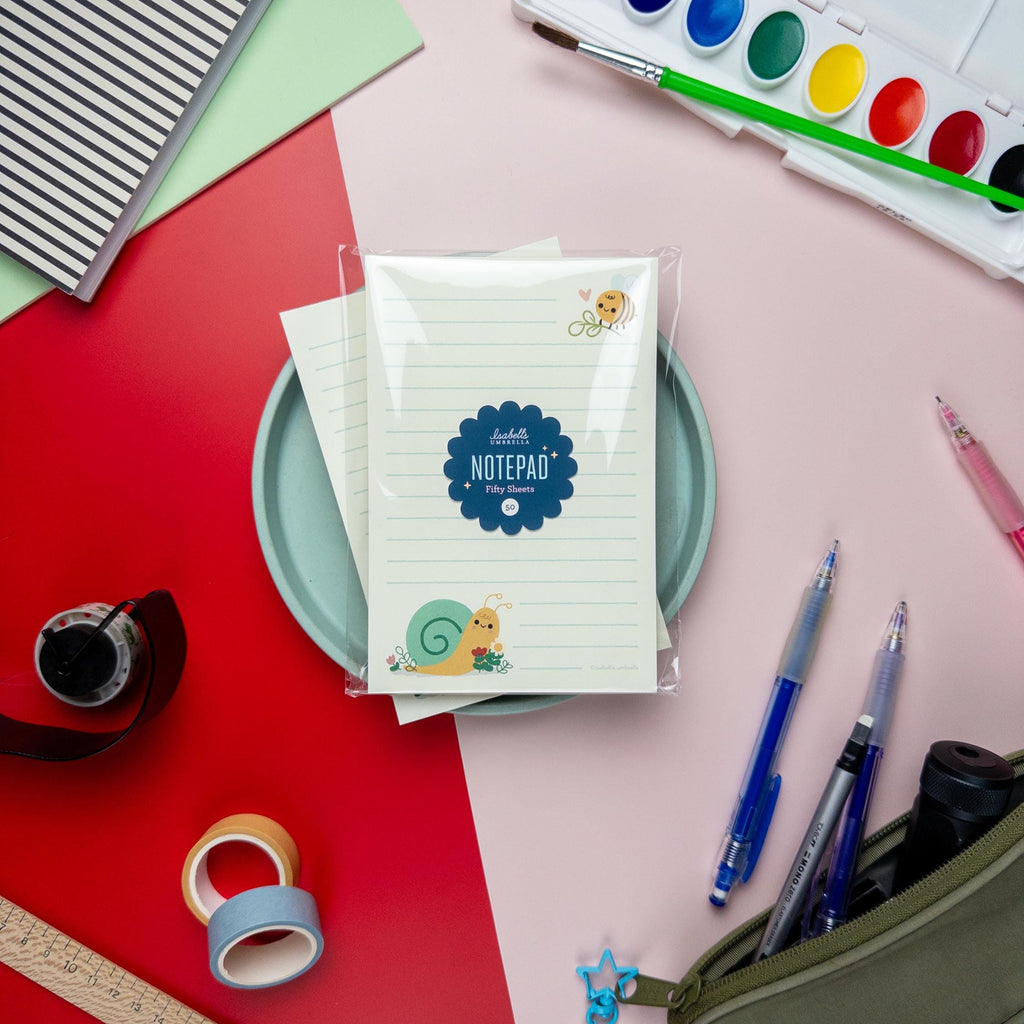 Snail and bee-themed notepad from Isabell’s Umbrella unwrapped and displayed on a pastel plate. The lined pages feature a smiling snail illustration at the bottom and a small bee with a heart at the top, surrounded by colorful art supplies.