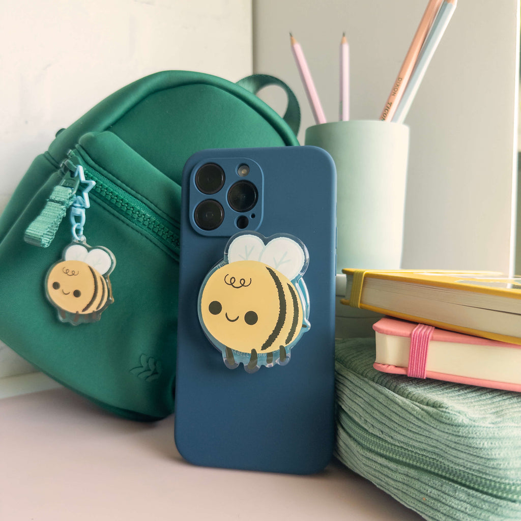Phone case with a yellow bee magnetic phone grip next to a green bag and books on a desk.