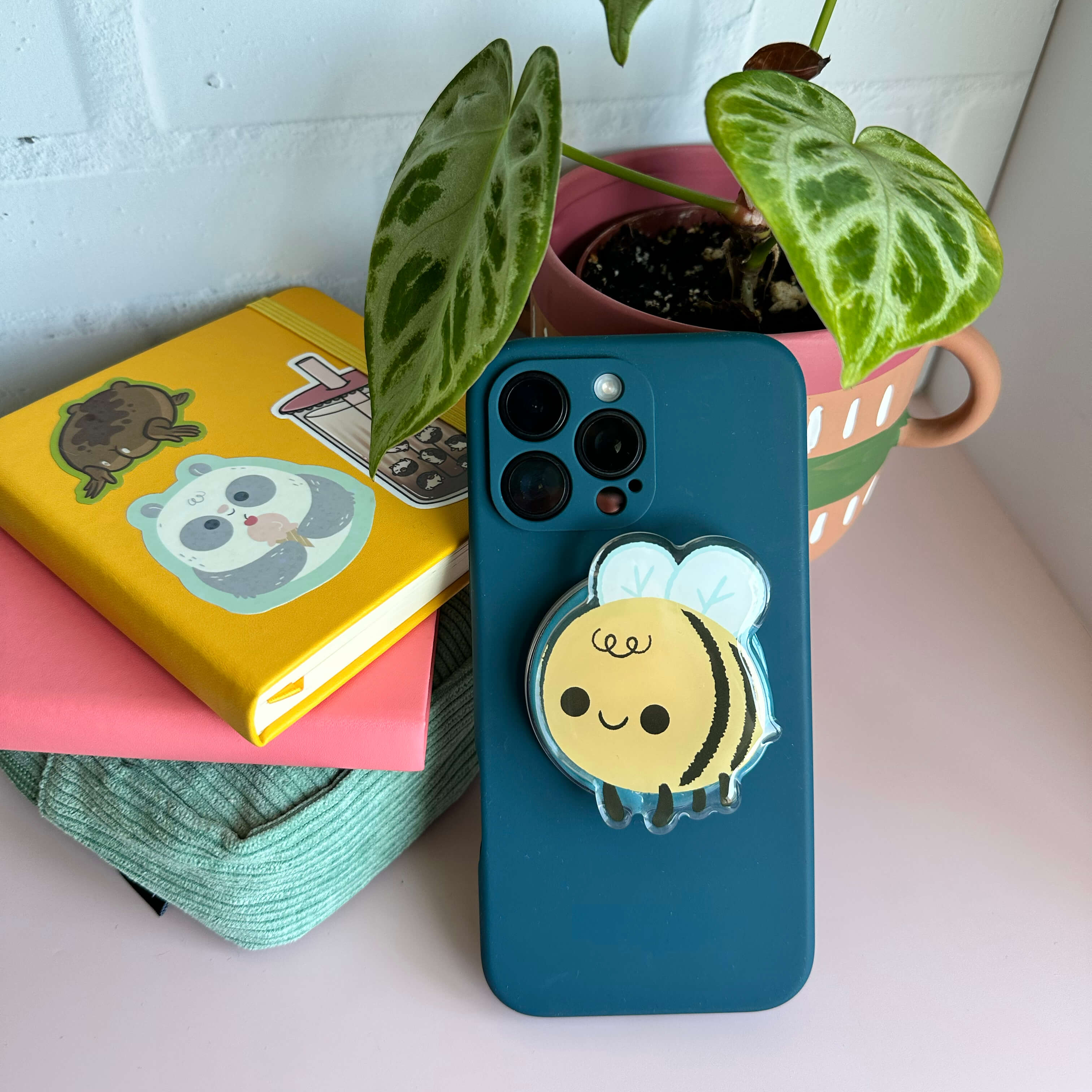 Phone case with a bee designed magnetic phone grip on a table with books and a plant
