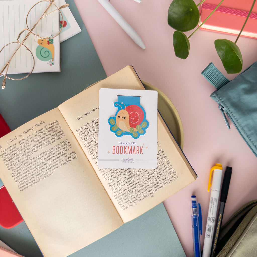 Magnetic bookmark with a yellow snail design on an open book surrounded by stationery items.