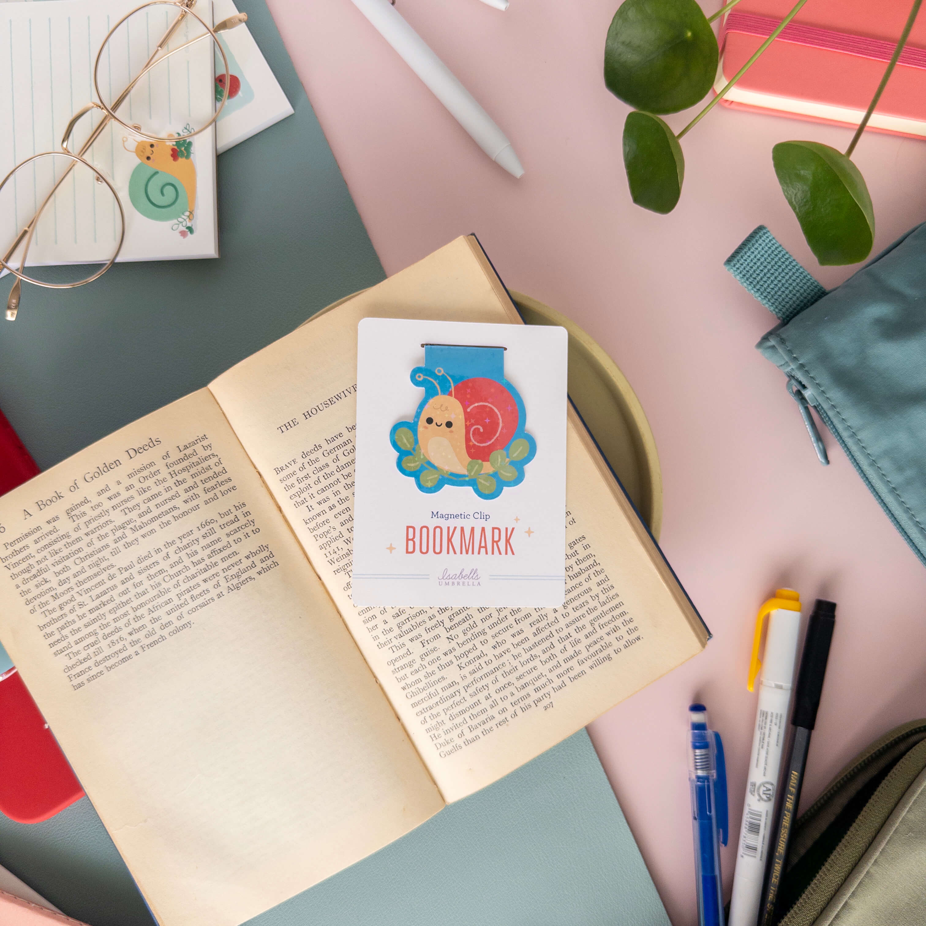 Magnetic bookmark with a yellow snail design on an open book surrounded by stationery items.
