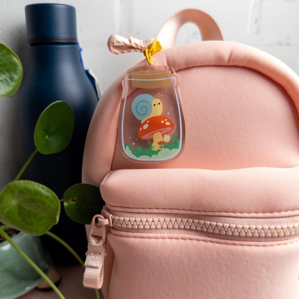 Pink backpack with a keychain of a snail in a terrarium jar. It has been attached to zipper pull and it pictured beside a blue water bottle and small plant.