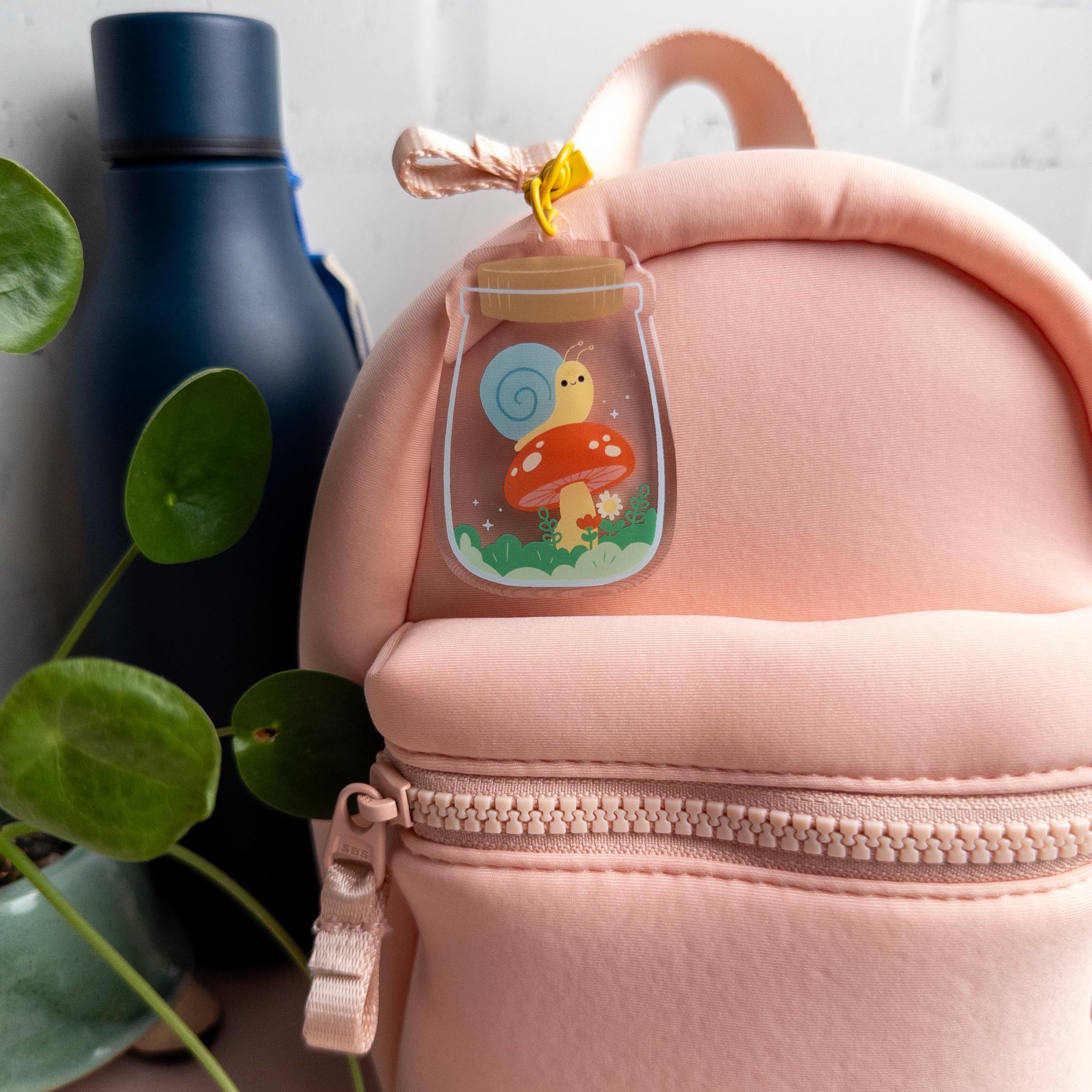Pink backpack with a keychain of a snail in a terrarium jar. It has been attached to zipper pull and it pictured beside a blue water bottle and small plant.