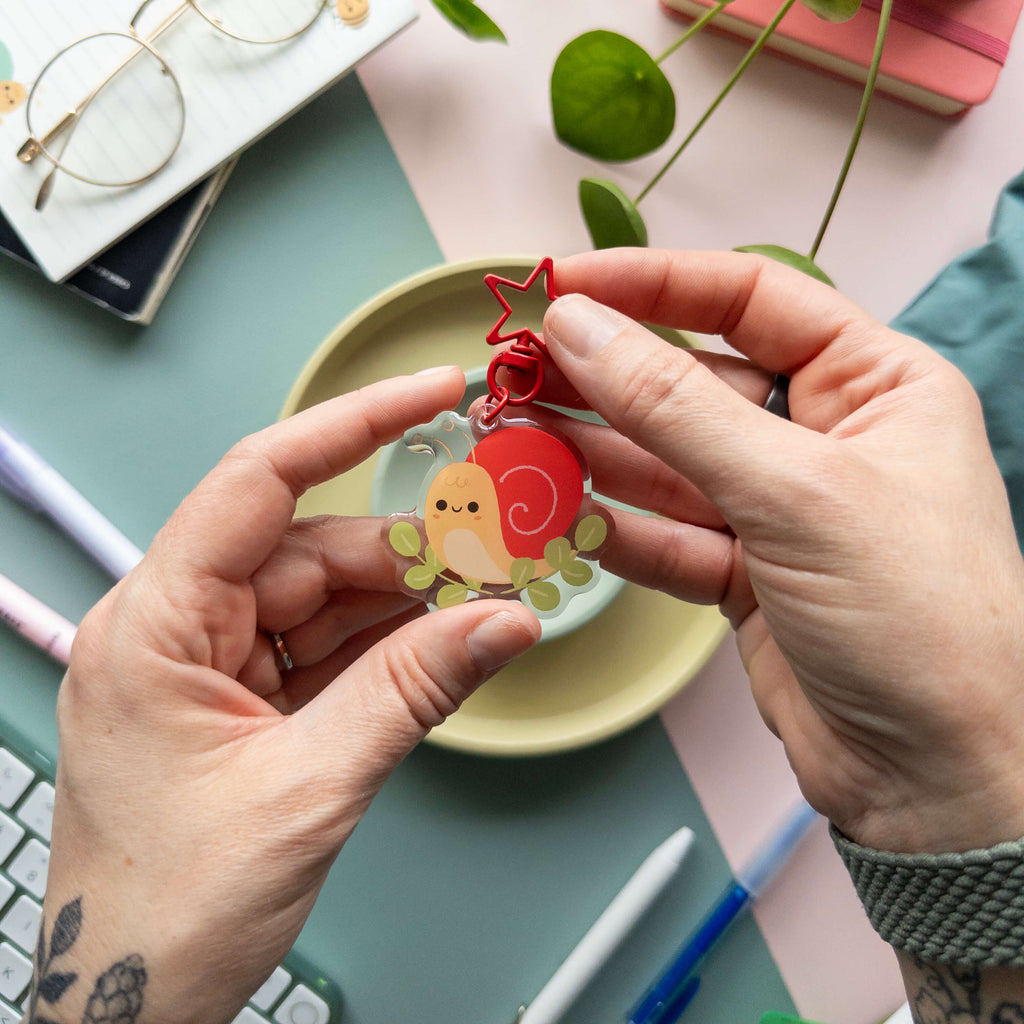 Person holding a yellow snail charm with a red shell over  a desk with stationery items.
