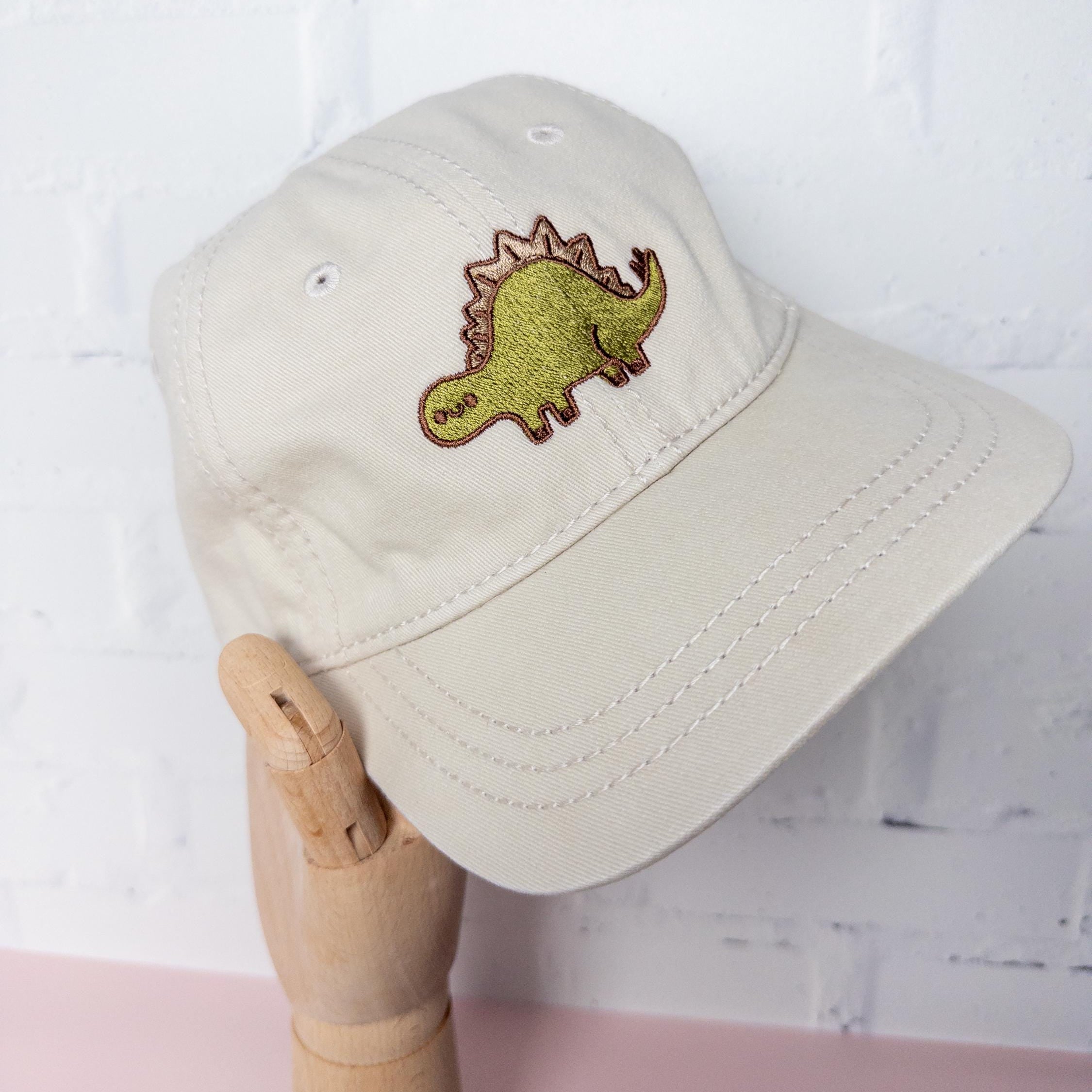 Wood model hand holding a cream cotton twill dad hat with embroidered green stegosaurus design, adjustable dinosaur baseball cap for dino lovers and minimalist fashion fans.