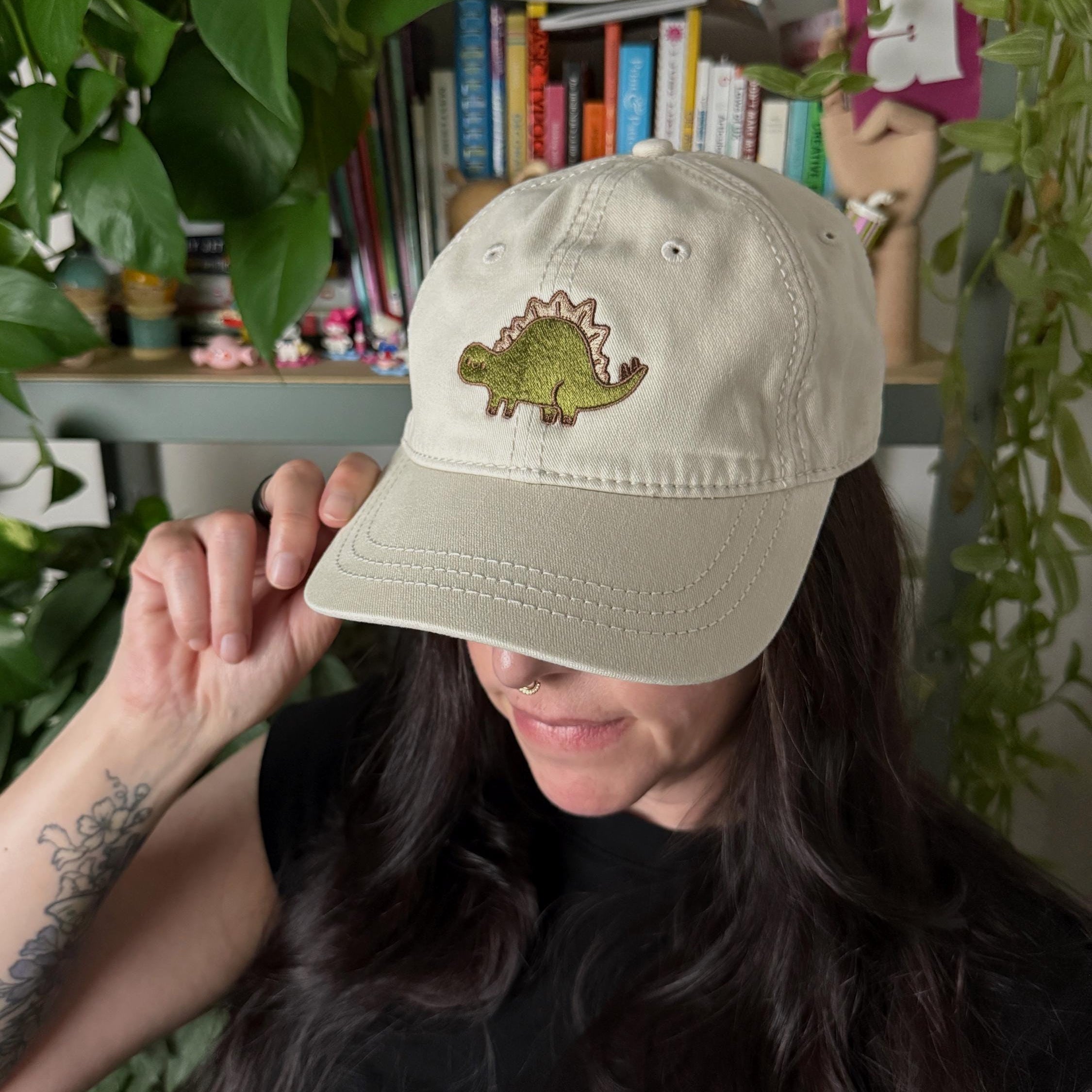Woman wearing a cream cotton twill dad hat with embroidered green stegosaurus design.
