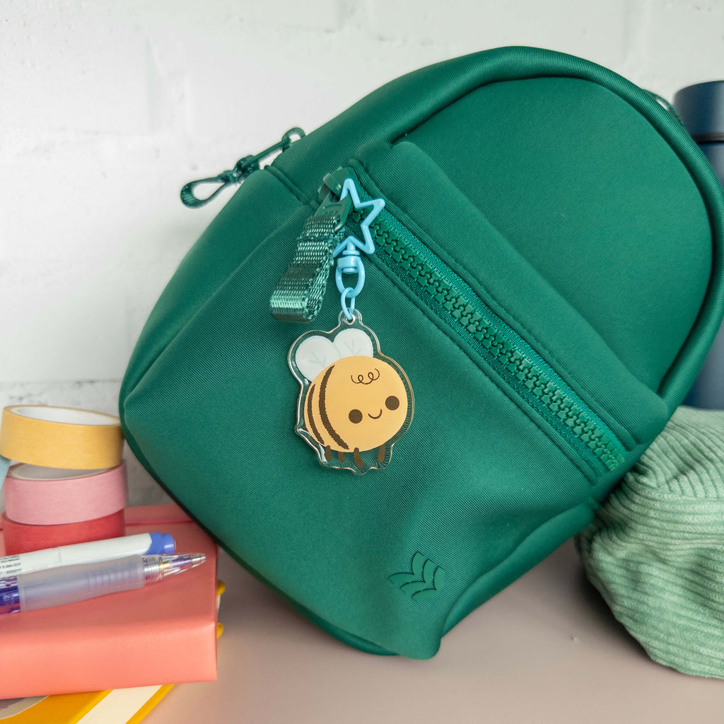 Green small backpack with a bee charm keychain on a surface with stationery items