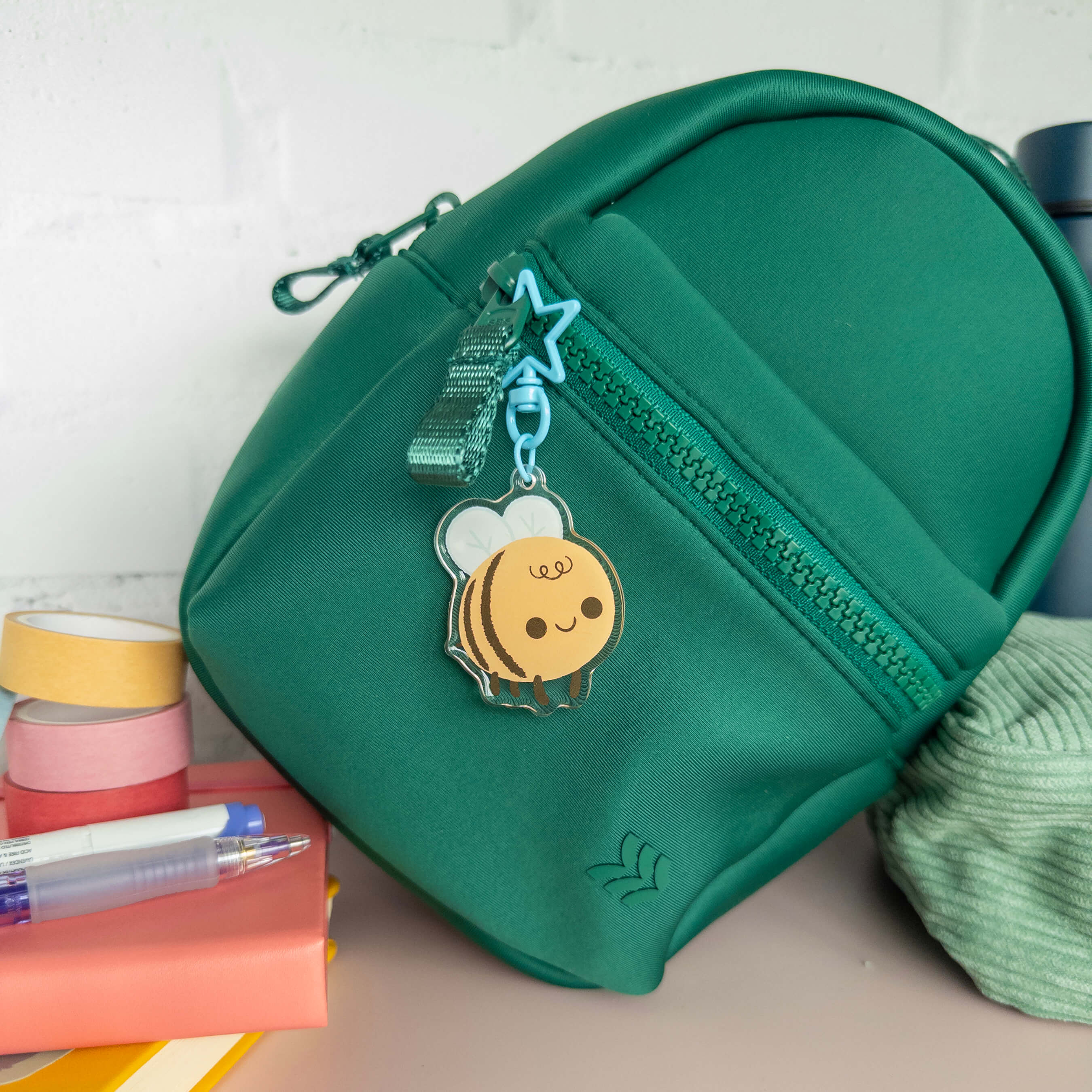 Green small backpack with a bee charm keychain on a surface with stationery items