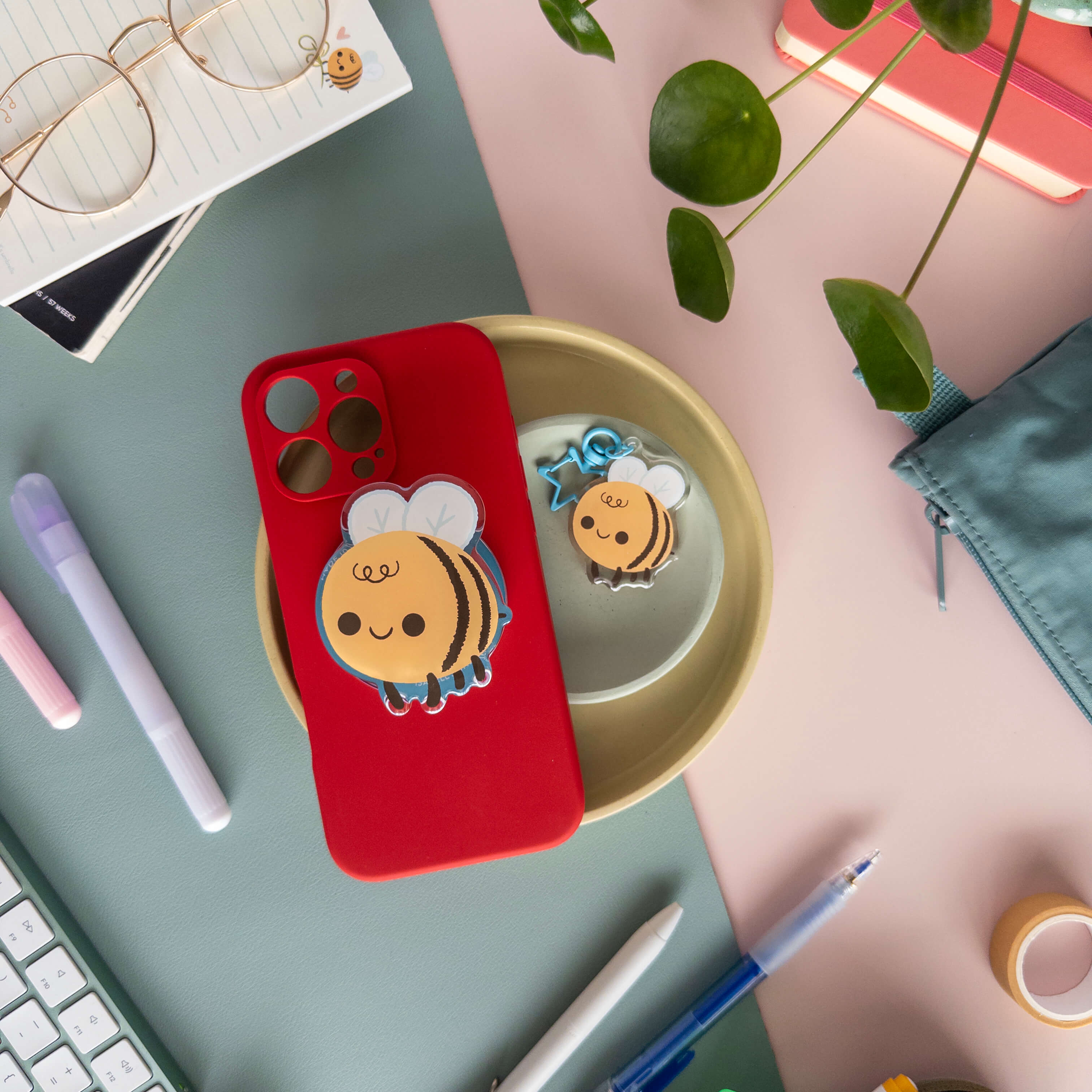 Red phone case with a bee design magnetic phone grip next to a acrylic bee keychain on a desk with stationery items