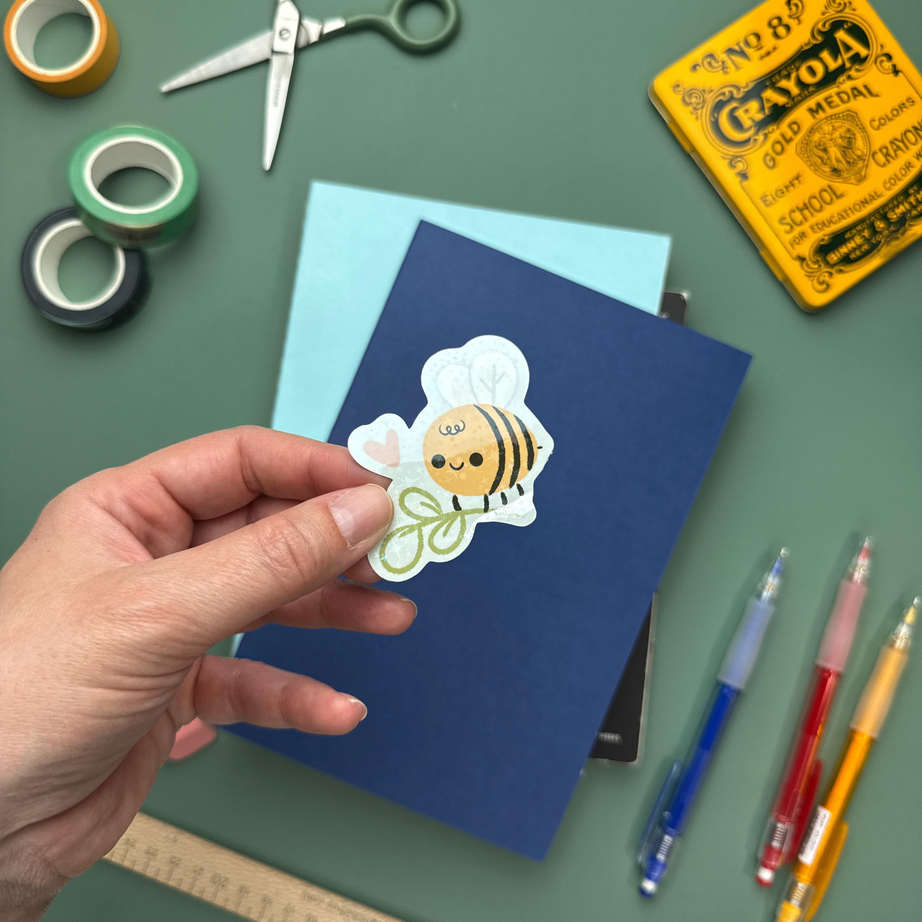 Cute glitter bee vinyl sticker with sparkly finish, waterproof and durable decal for laptops, water bottles, and notebooks held by a hand with journals and other art supplies in the background.